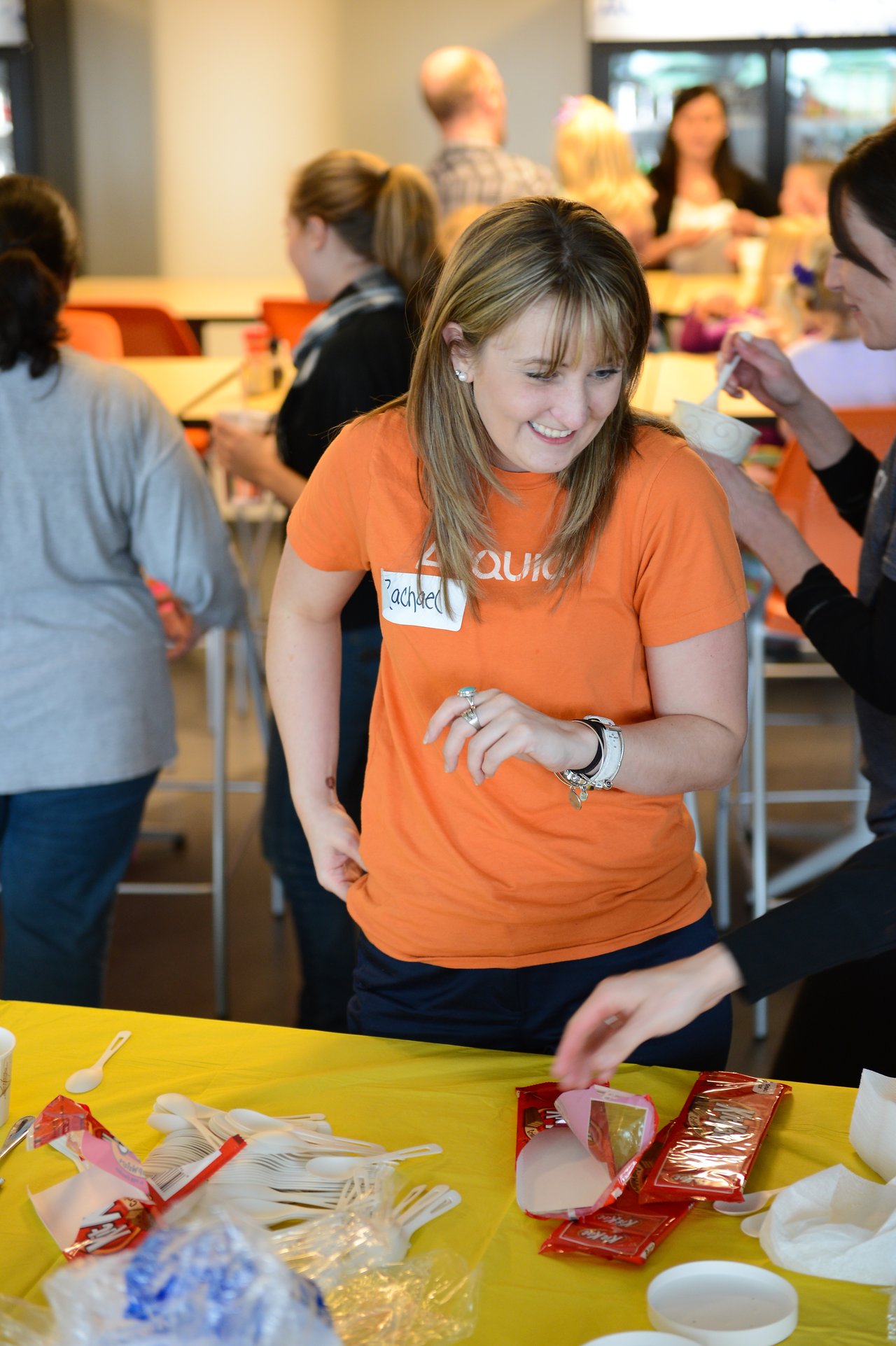 A woman in an orange shirt smiles while standing near a table with plastic utensils and food items.