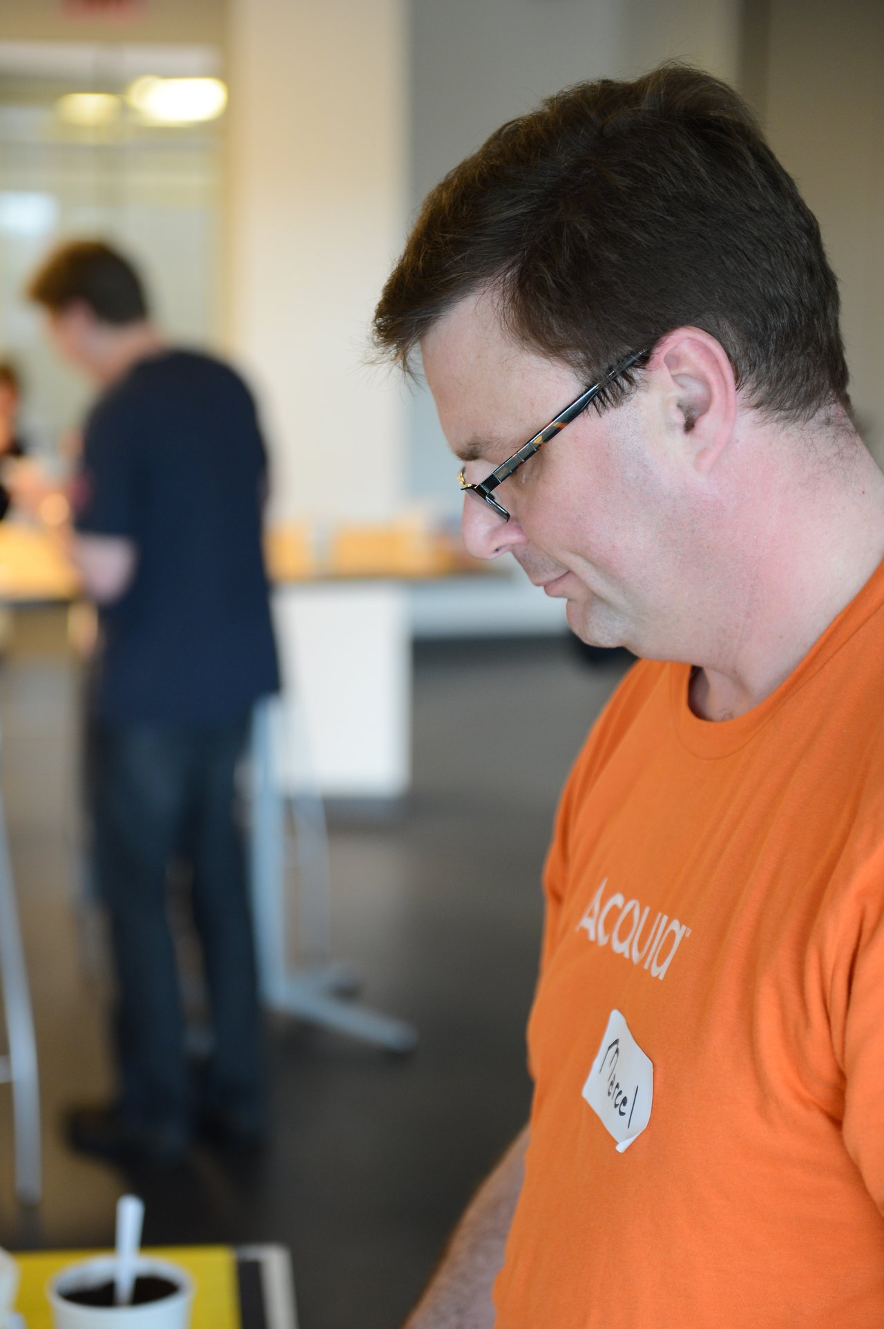 A man in an orange Acquia shirt looks down while standing indoors.