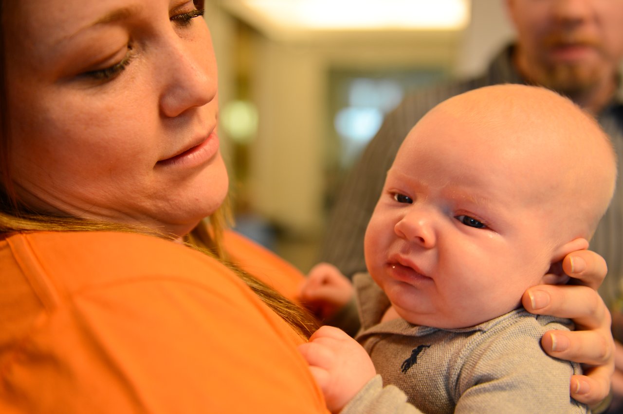A woman in an orange shirt holds a baby, who appears to be fussy or uncomfortable.