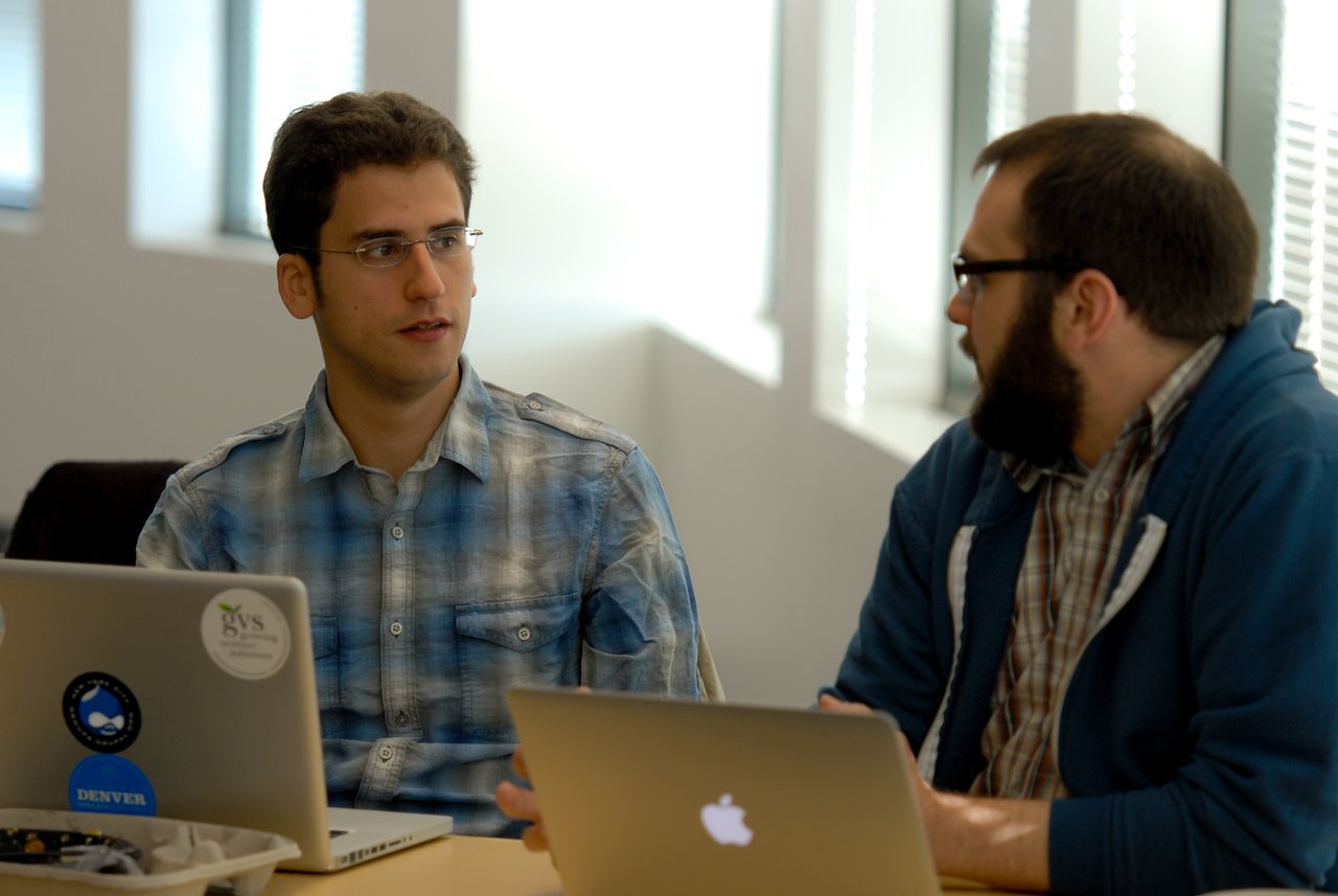 Two people sit at a table with laptops, engaged in a discussion in a well-lit office setting.