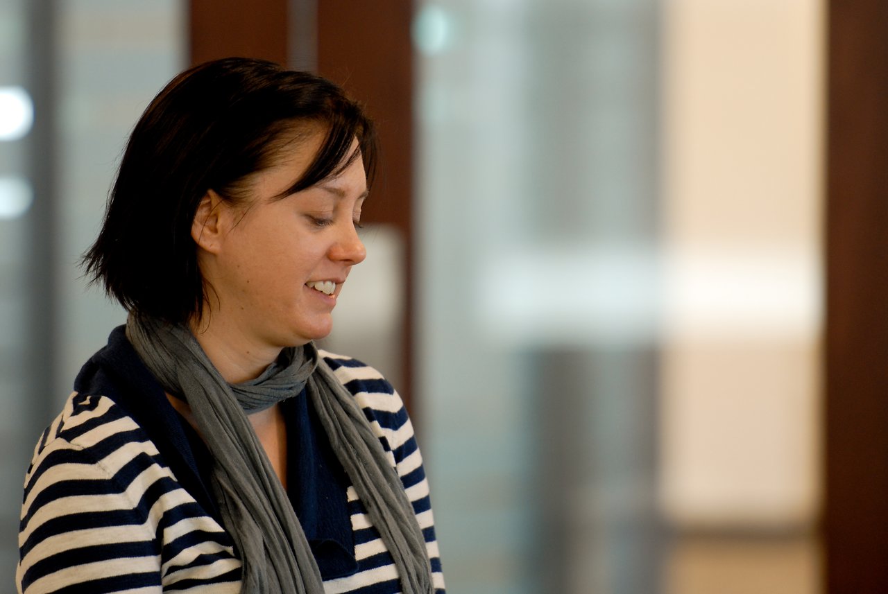 A person wearing a striped sweater and scarf is smiling and looking downward in an indoor setting.
