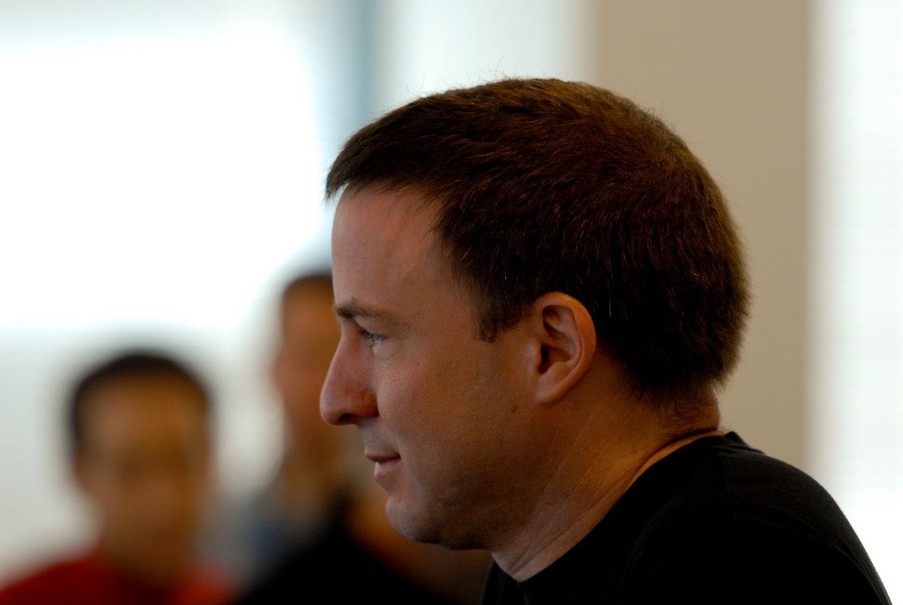 A man in a black shirt listens attentively, with blurred people in the background.