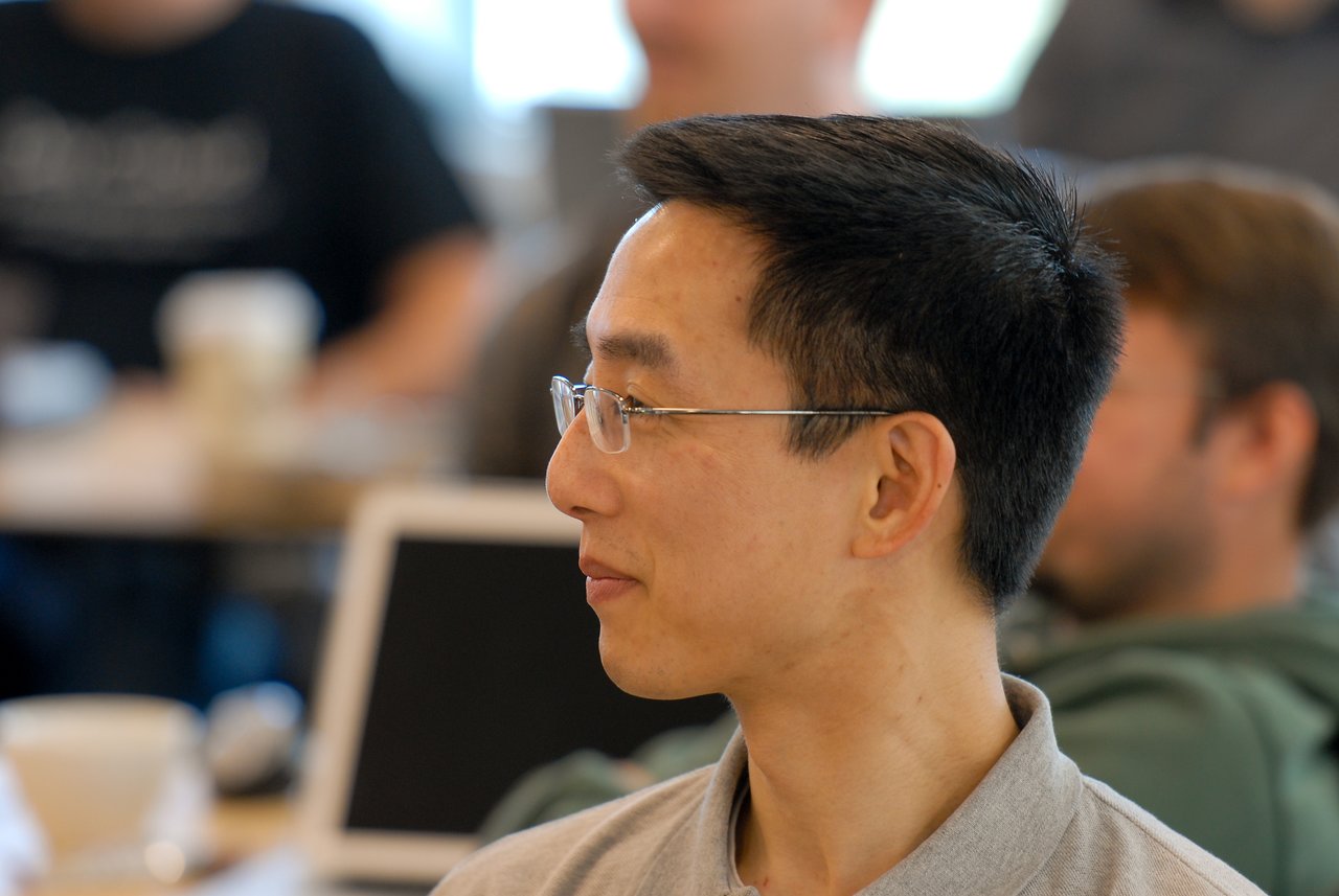 A man wearing glasses attentively listens during a meeting or conference, with others and laptops in the background.