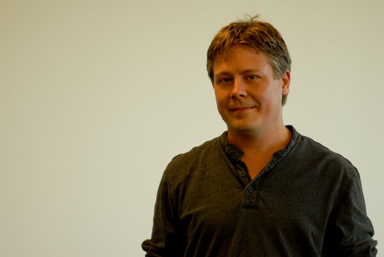 A person in a dark shirt stands in front of a plain background, looking at the camera with a slight smile.
