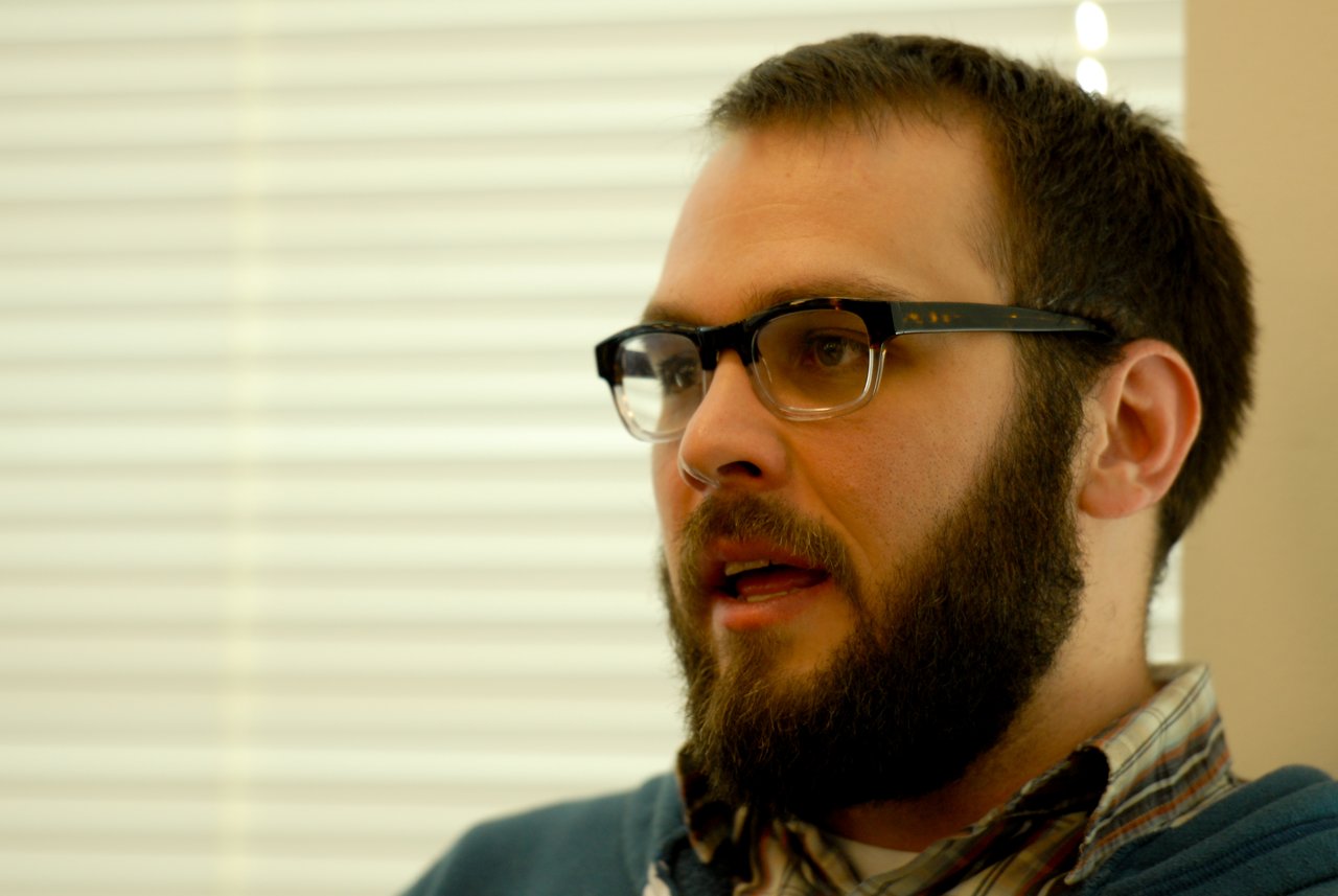 A bearded man wearing glasses speaks while looking to the side in an indoor setting.