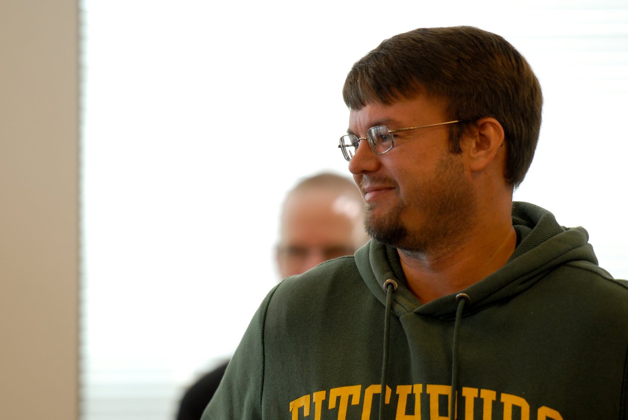 A man in a green hoodie and glasses smiles while looking to the side, with another person blurred behind him.