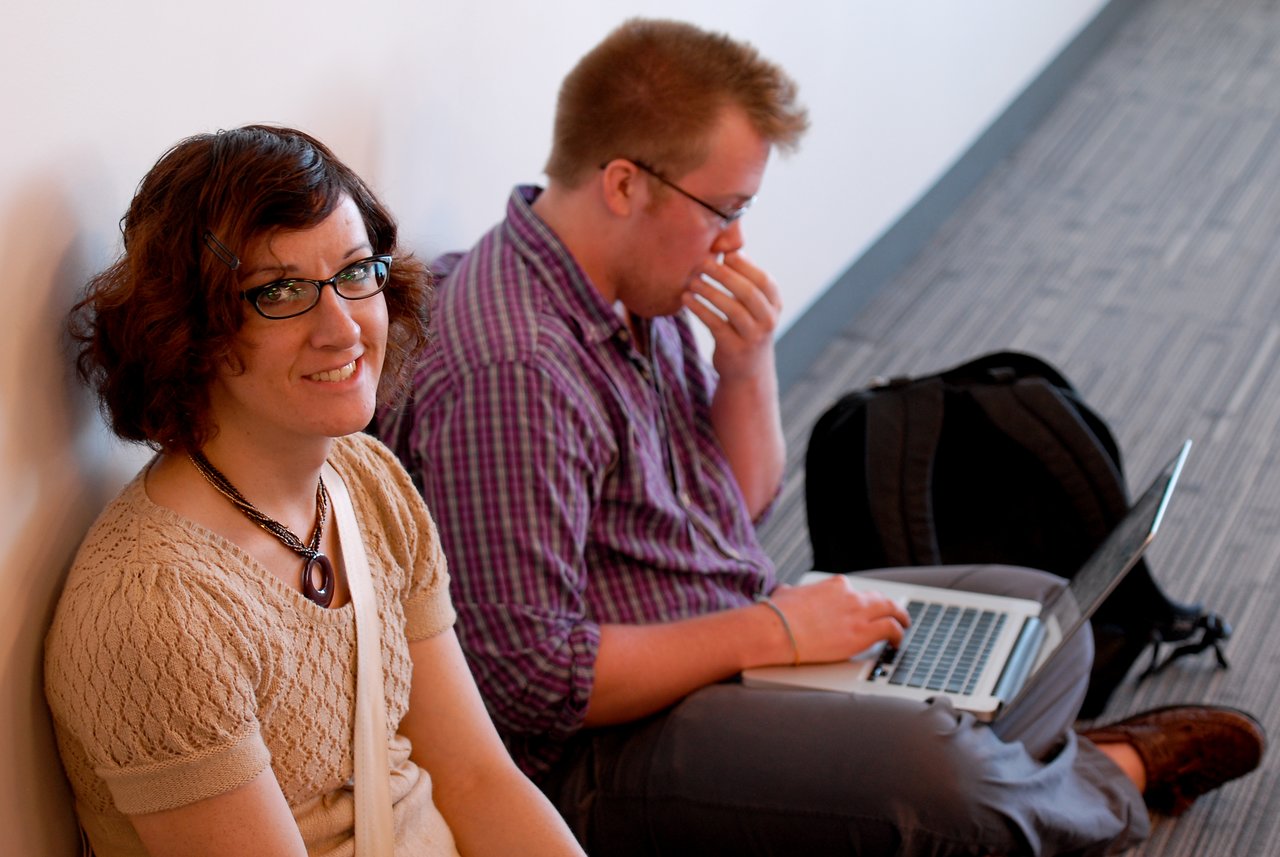 A woman smiles at the camera while a man sits beside her, focused on his laptop.
