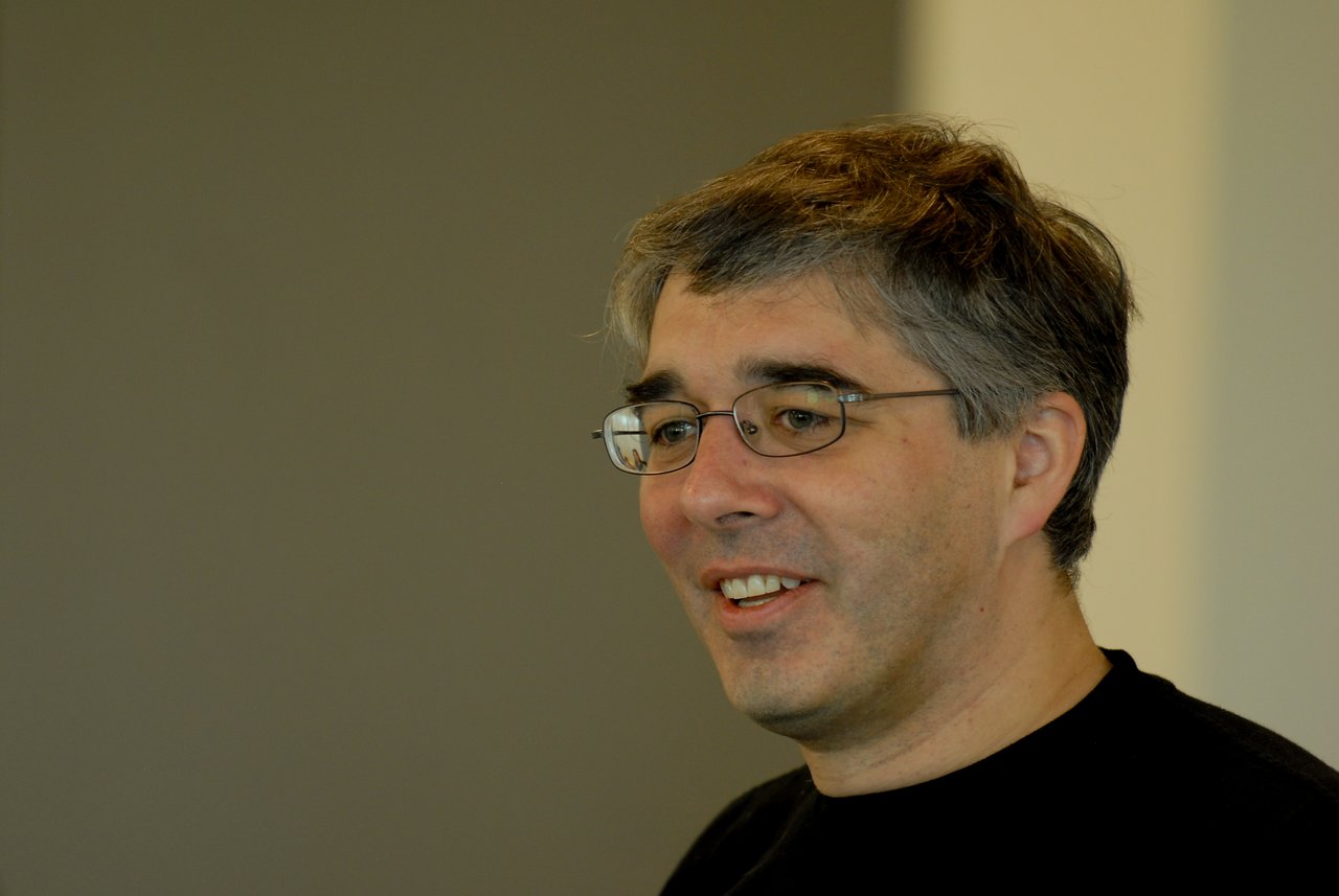 A man wearing glasses and a black shirt is speaking and smiling in an indoor setting.