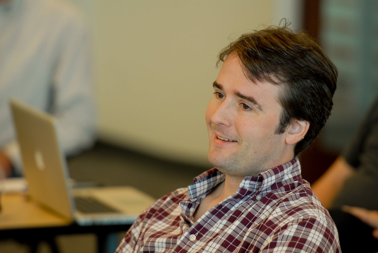 A man in a plaid shirt smiles while listening during a meeting or discussion.