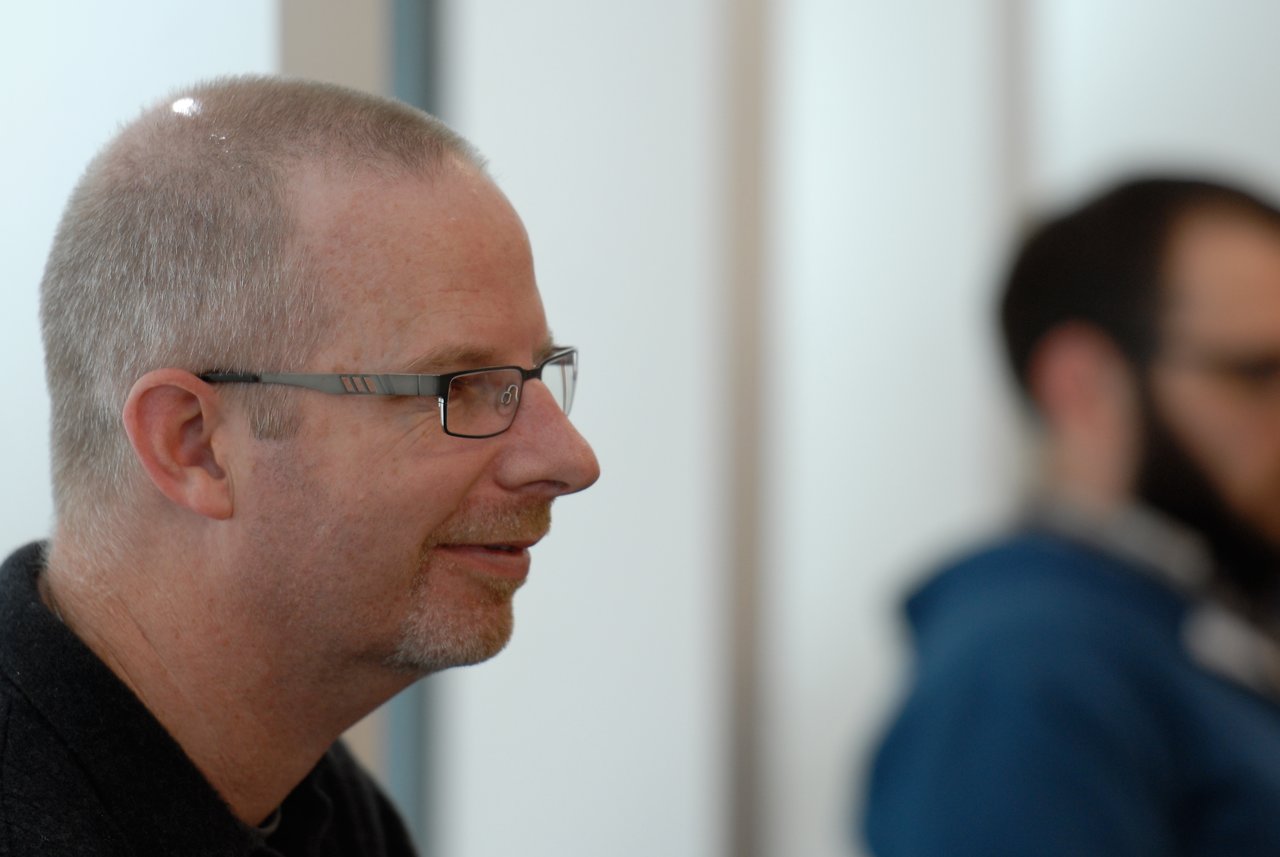 A man wearing glasses smiles while listening during a discussion, with another person blurred in the background.