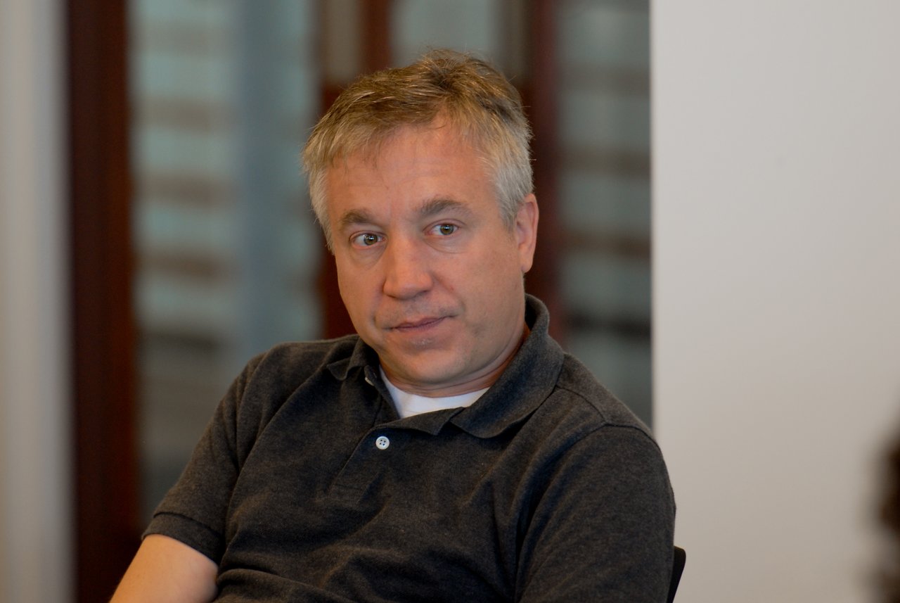 A man in a dark polo shirt sits indoors, looking to the side with a neutral expression.