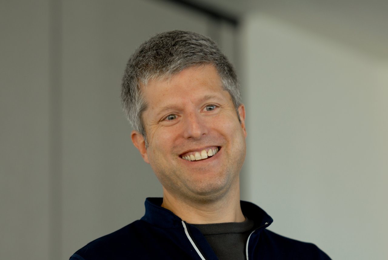 A smiling person with short gray hair wearing a dark jacket, looking slightly off-camera in an indoor setting.