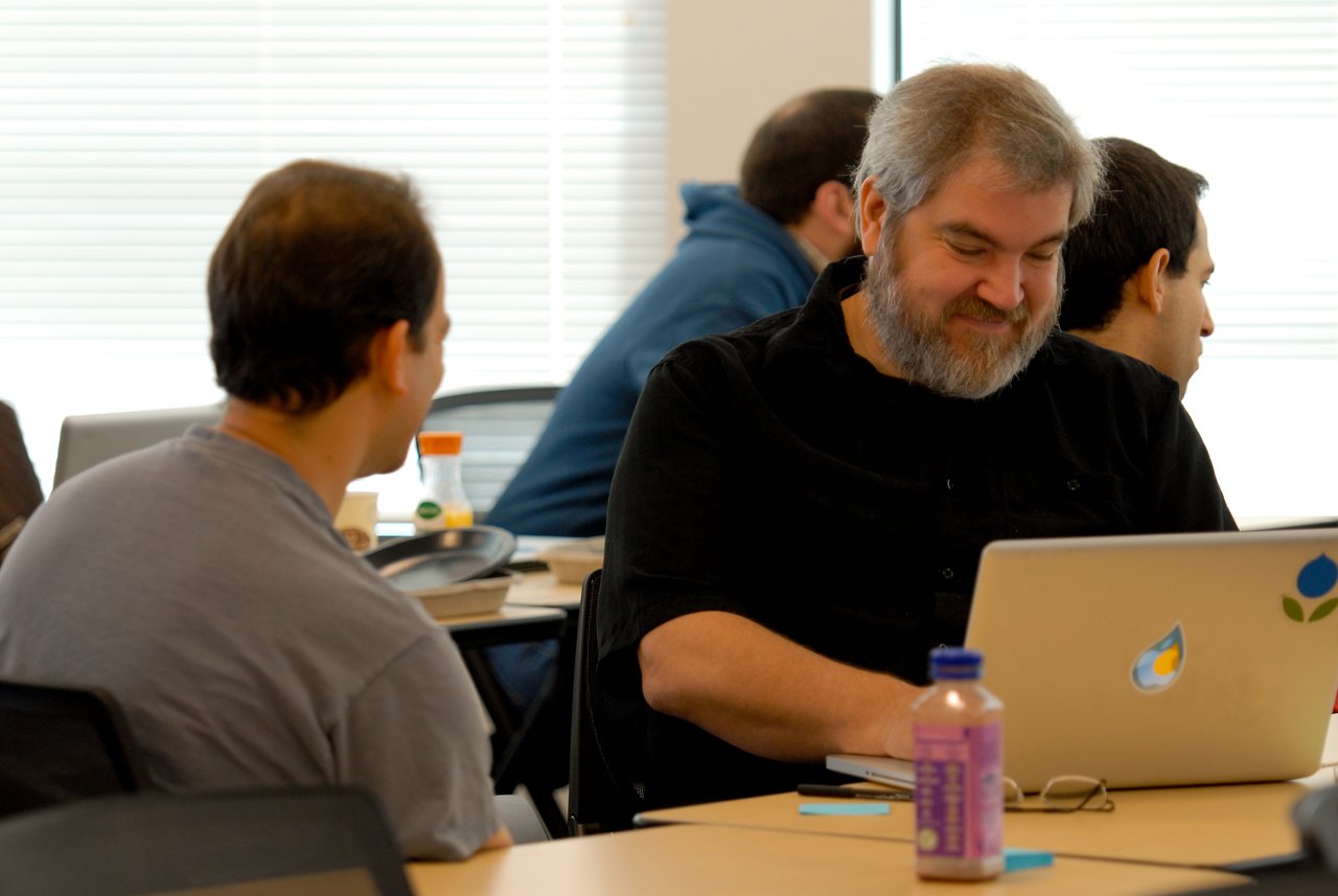 A bearded man smiles while working on a laptop, as another person sits nearby and talks to him.