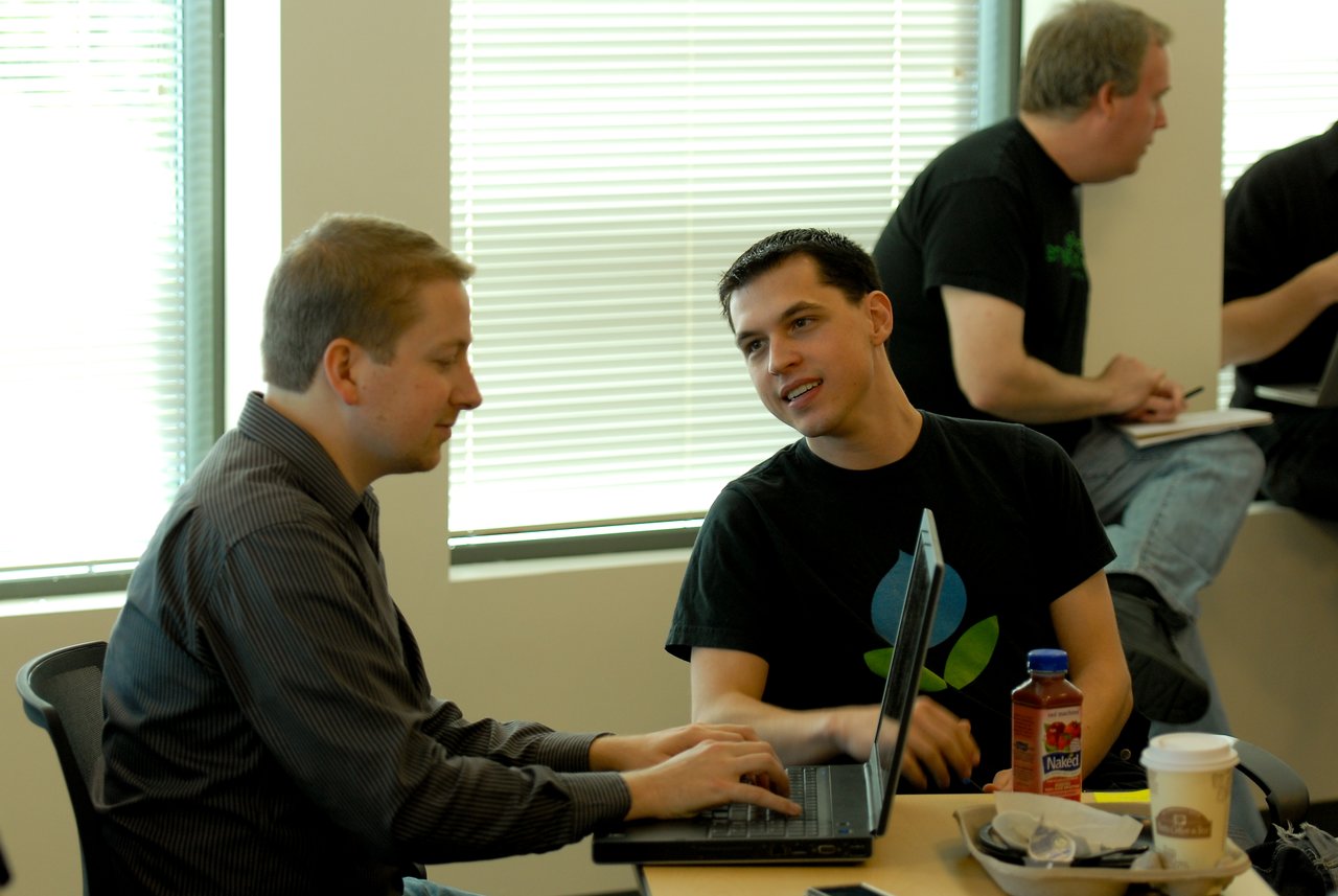 Two people sit at a table, discussing something on a laptop, while others work in the background.