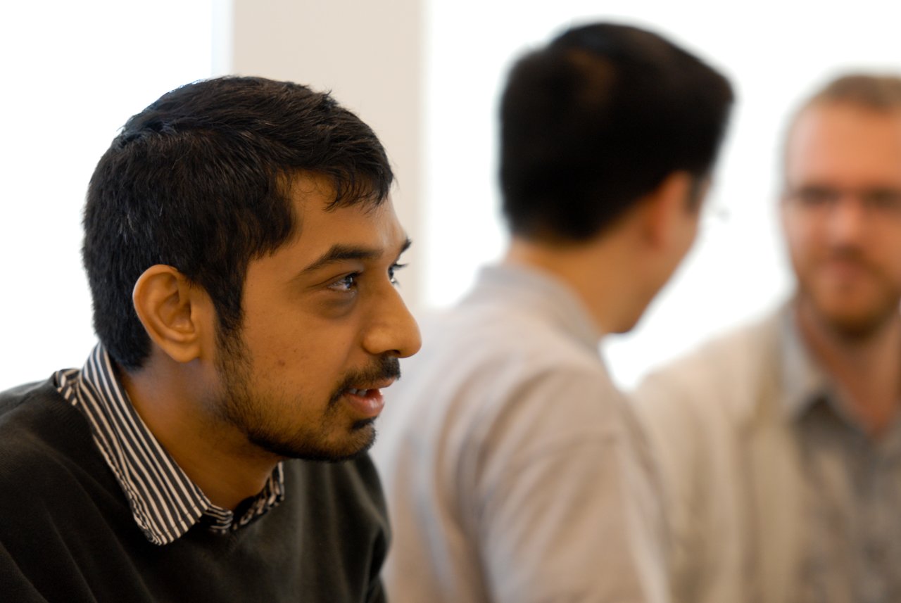 A man in the foreground speaks while two others in the background engage in conversation.