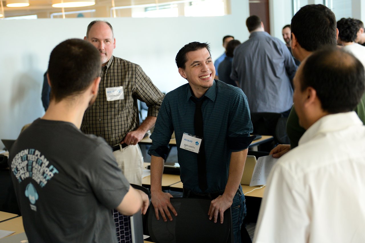 A group of people engaged in conversation at a professional event, with one person smiling and leaning on a chair.