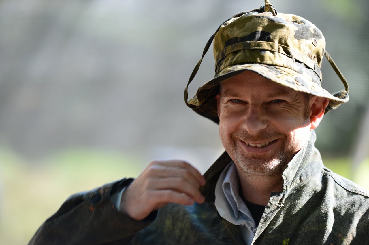 A smiling man in a camouflage hat and jacket adjusts his collar while looking at the camera.