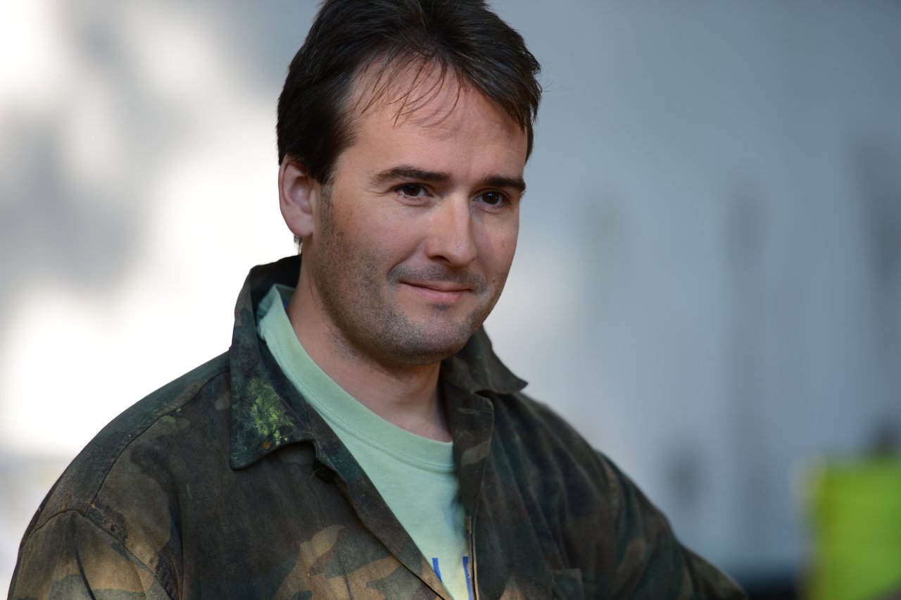 A person wearing a camouflage jacket and green shirt smiles slightly while looking off to the side.