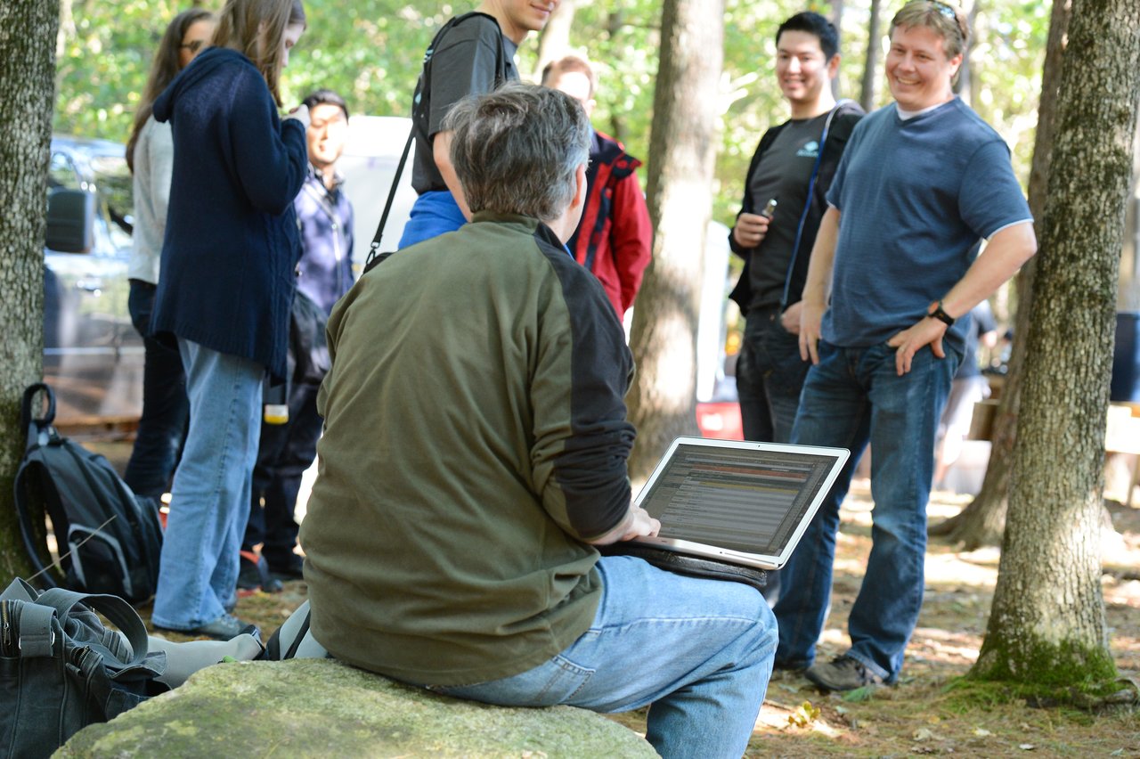 A person sits on a rock using a laptop while a group of people stands nearby, talking and smiling.