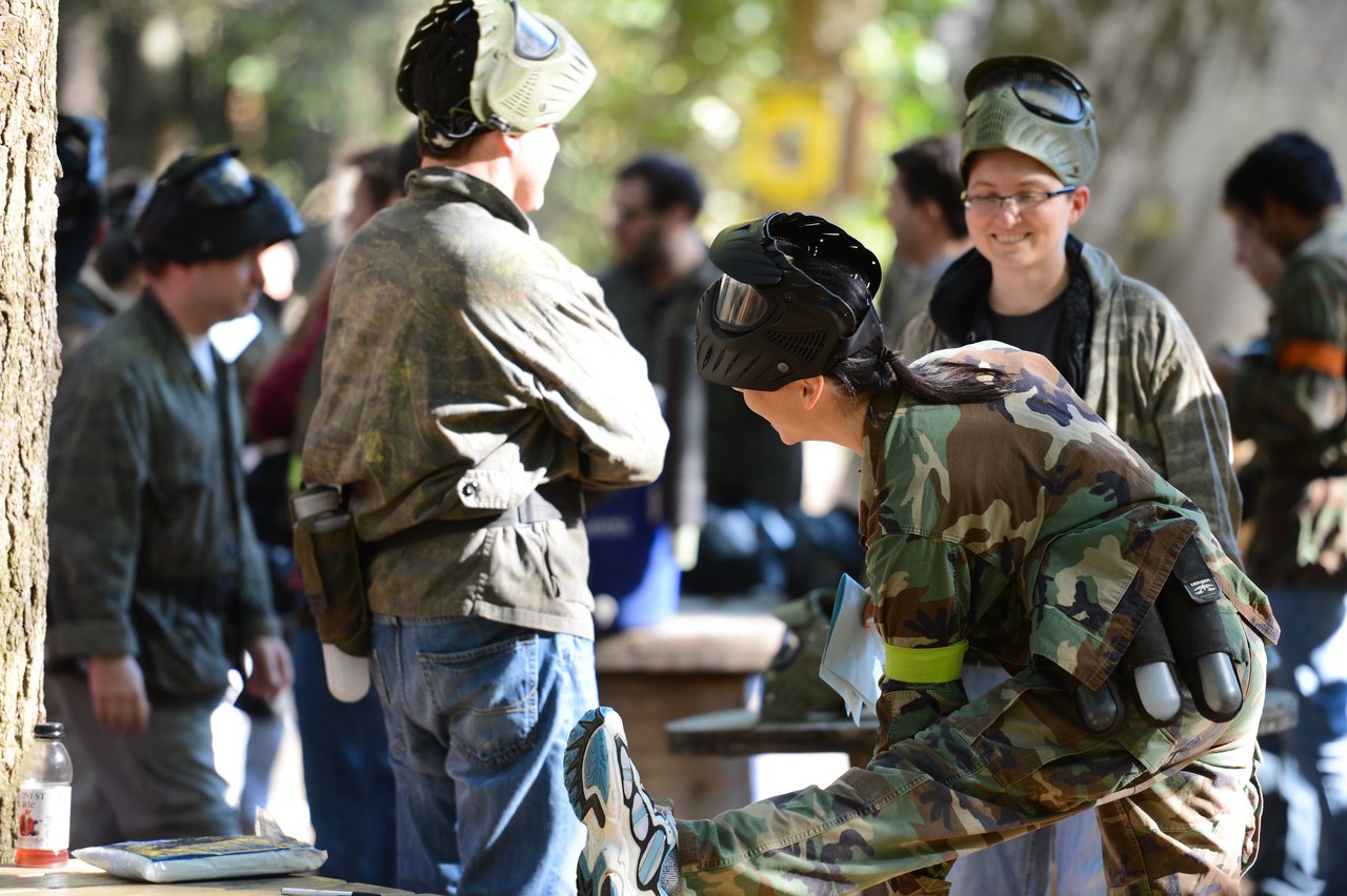 People in camouflage gear and protective masks prepare for a paintball game, with one person stretching and others chatting.