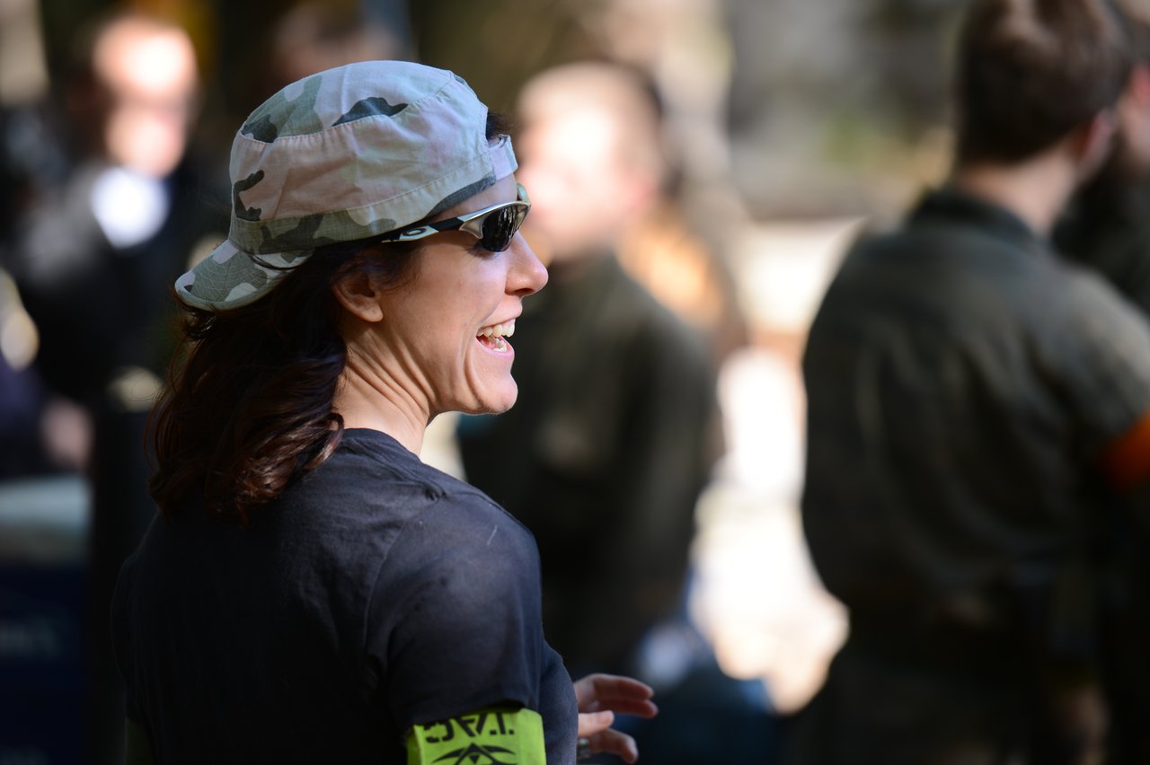 A person wearing a camouflage cap and sunglasses smiles while interacting with others in an outdoor setting.