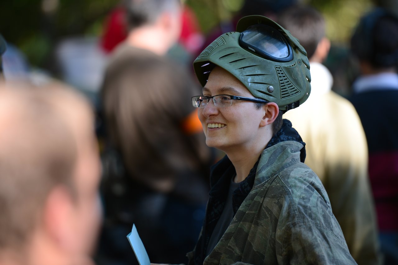 A person wearing a paintball mask and camouflage jacket smiles while holding a piece of paper in a crowded setting.