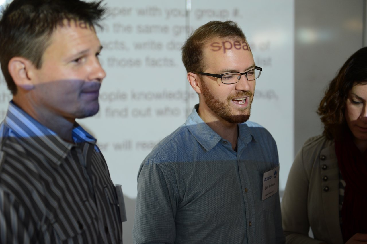 Three people stand in front of a projected screen, with one man speaking while the others listen.