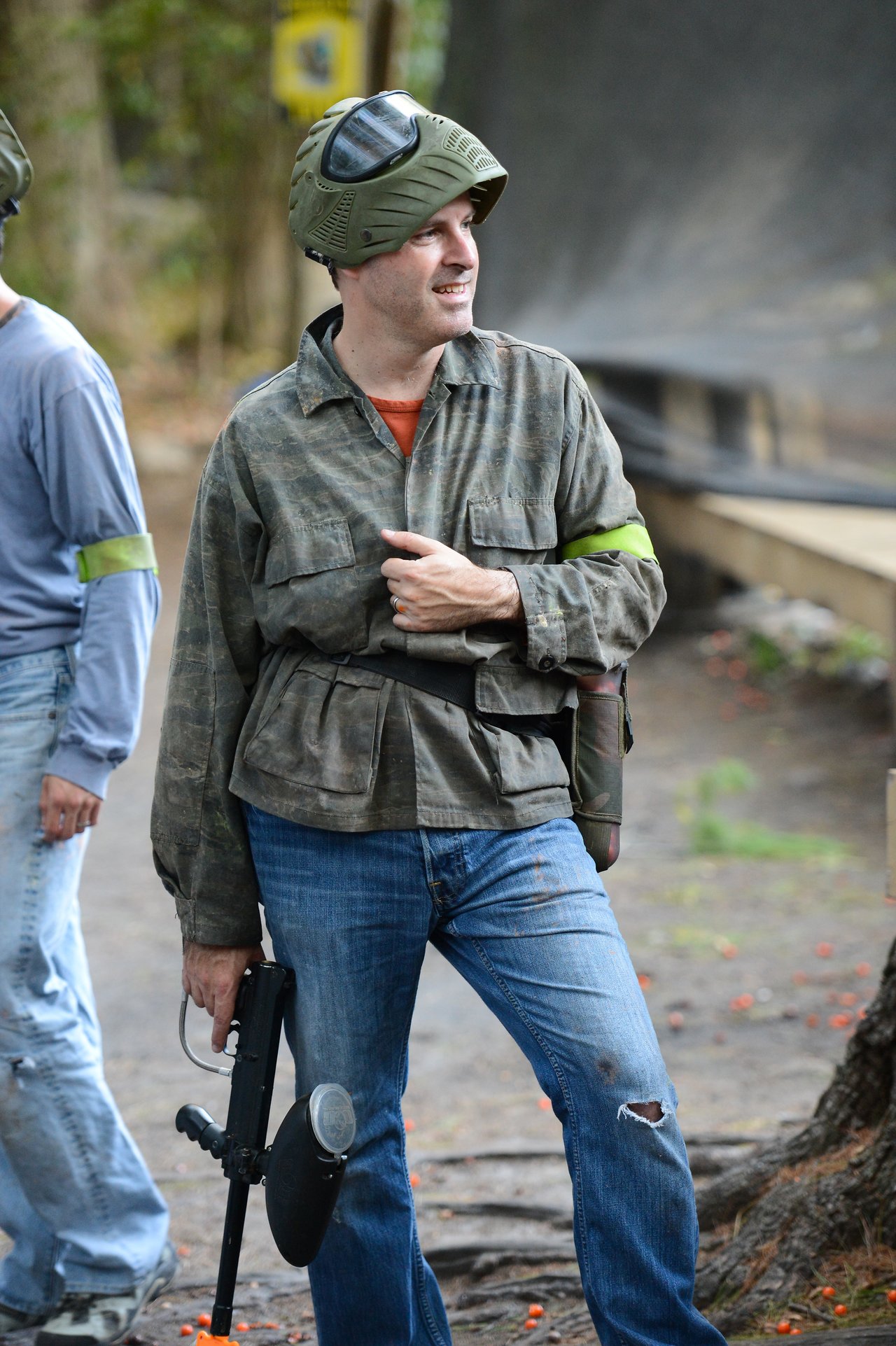 A person in paintball gear holds a paintball gun while standing outdoors, wearing a helmet and camouflage jacket.