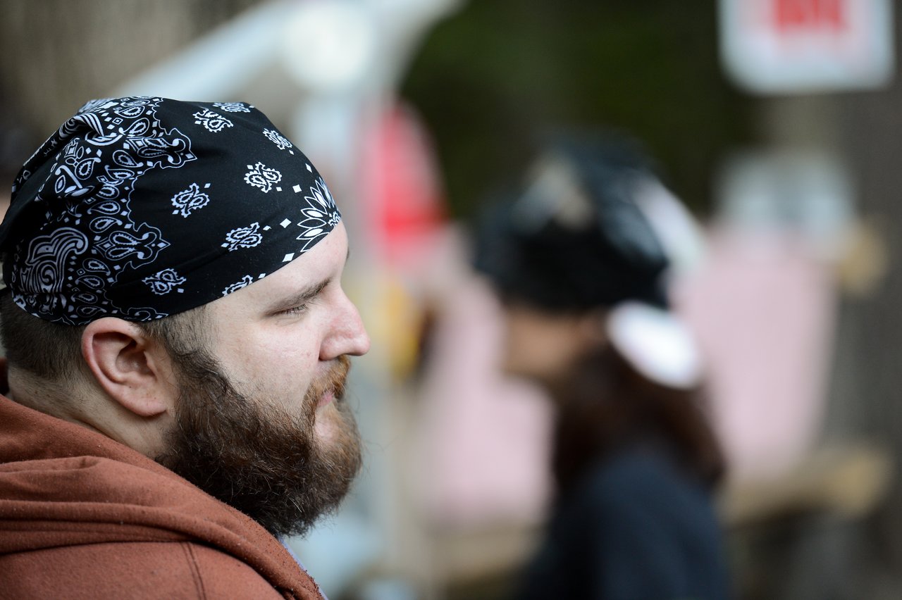 A bearded person wearing a black bandana looks to the side, with another person blurred in the background.