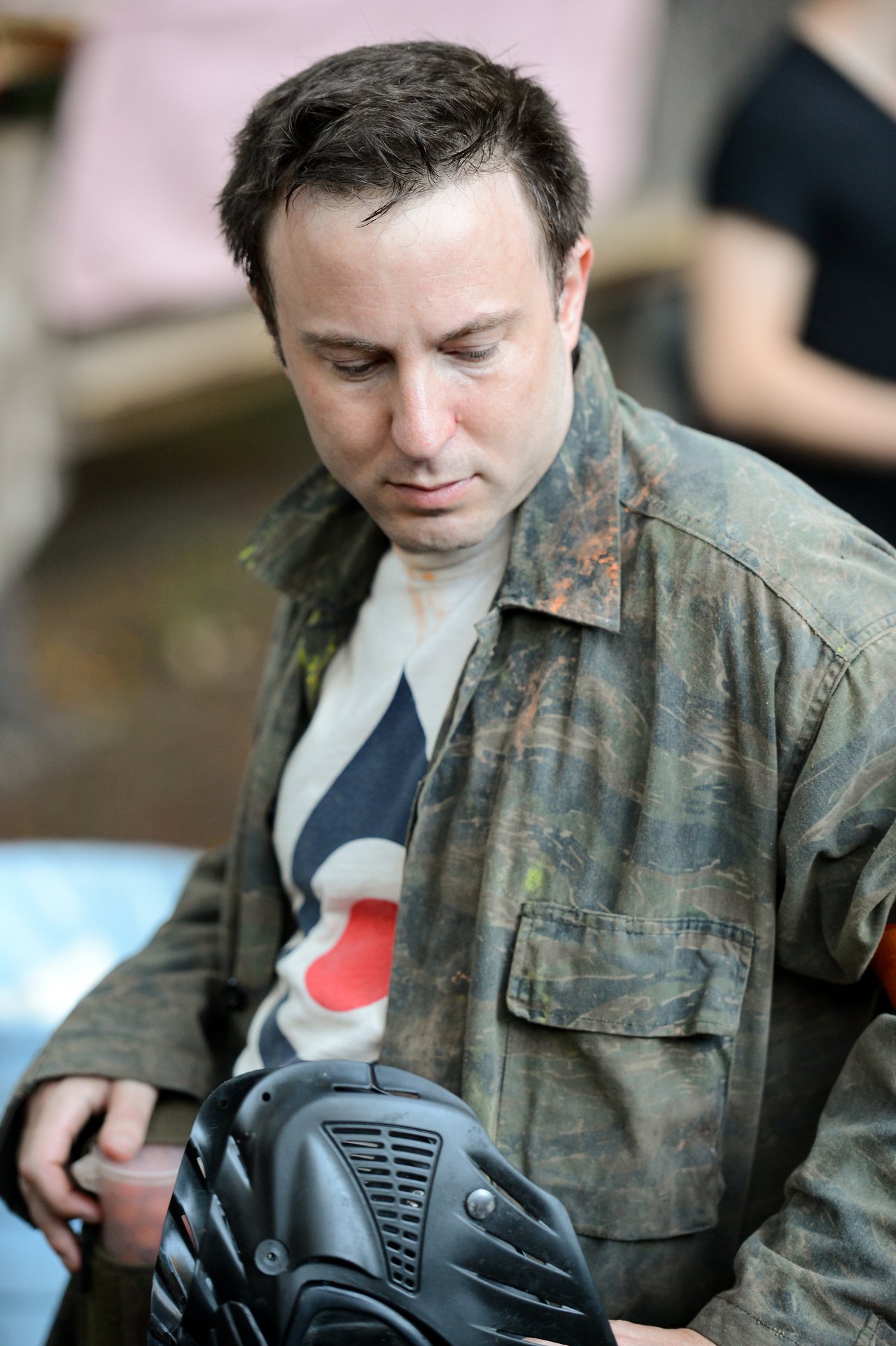 A person wearing a camouflage jacket holds a black paintball mask while looking down.