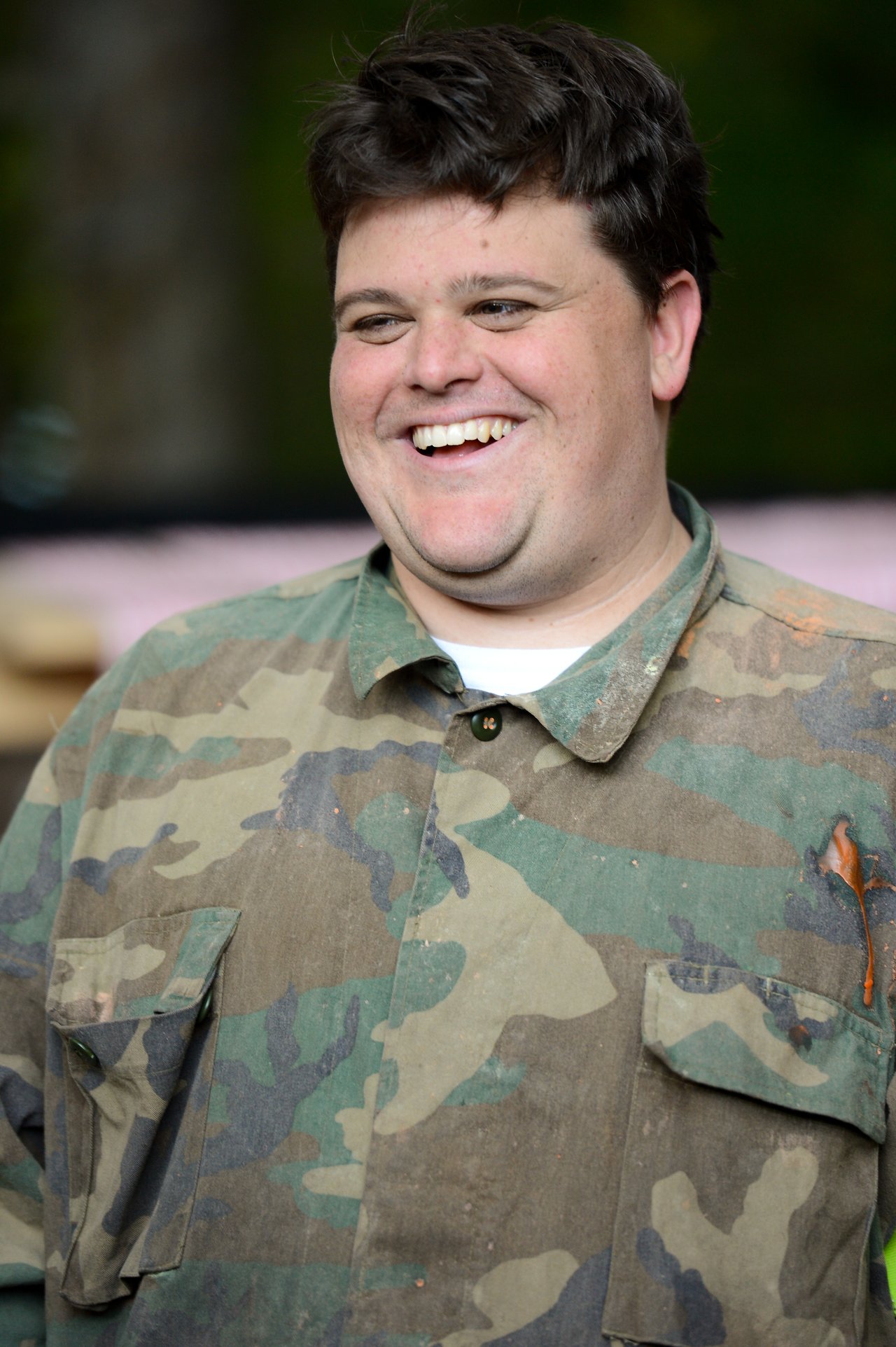 A smiling person wearing a camouflage shirt with some paint splatters on the fabric.