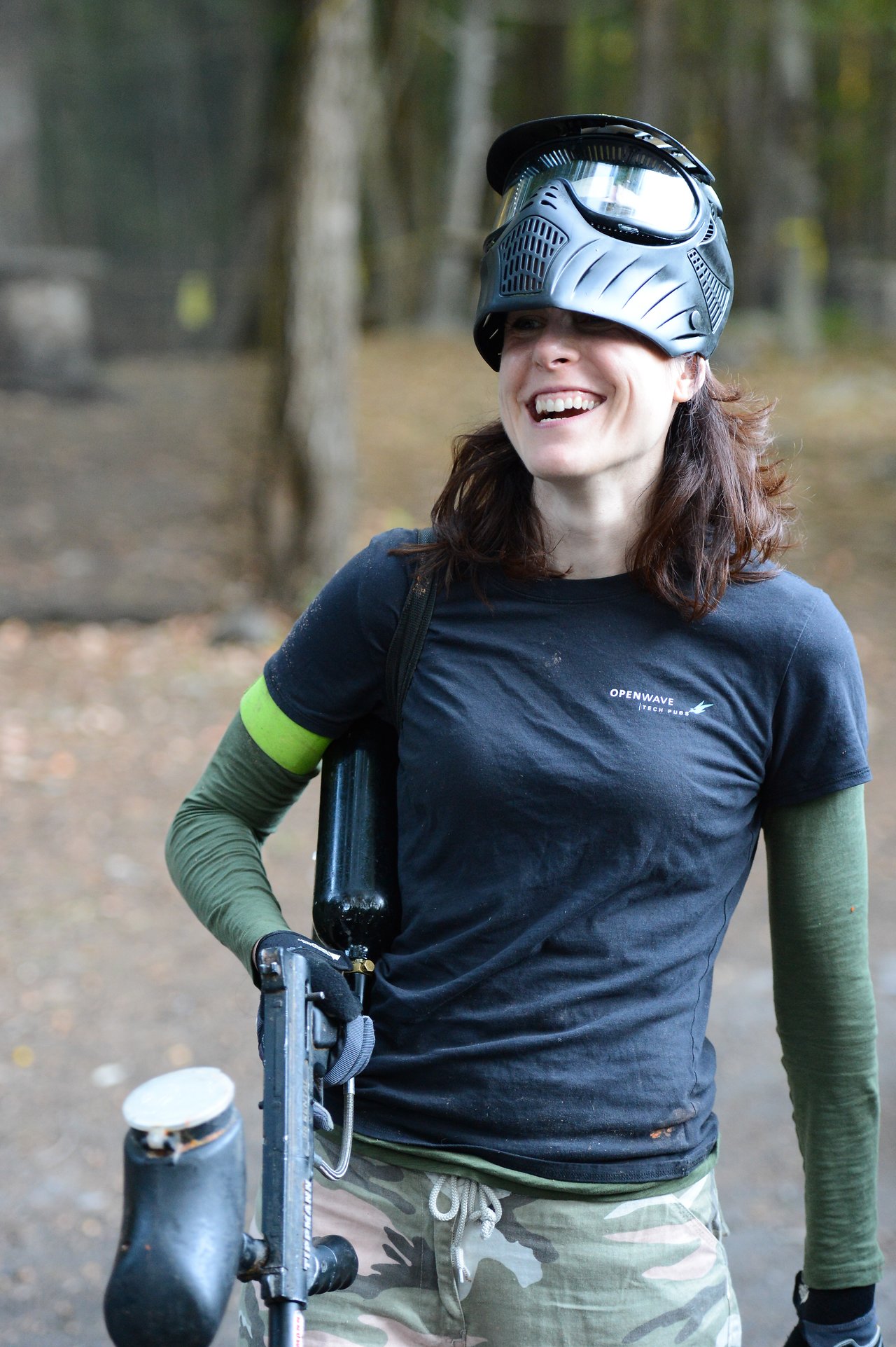 A person wearing a paintball mask and camouflage pants smiles while holding a paintball gun in an outdoor setting.