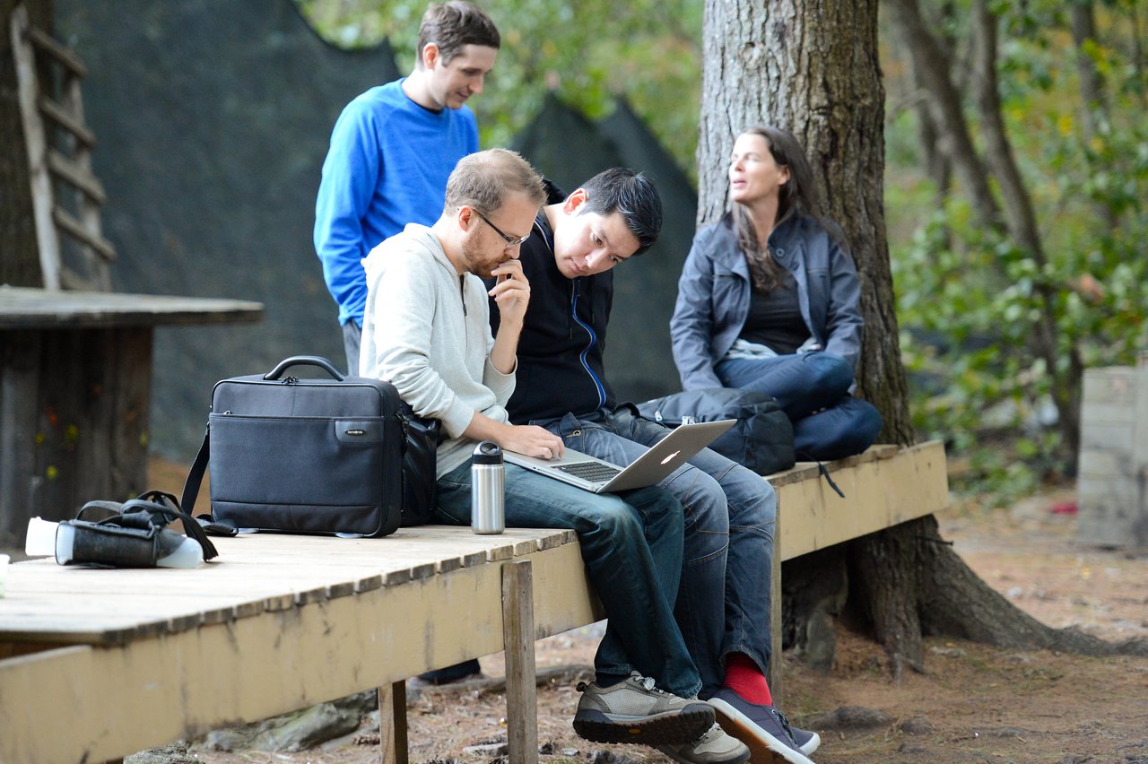 Two people sit on a bench, looking at a laptop, while two others sit and stand nearby in conversation.