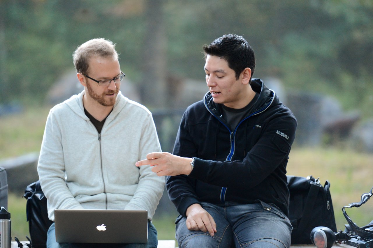 Two people sitting outdoors, one using a laptop while the other gestures and speaks.