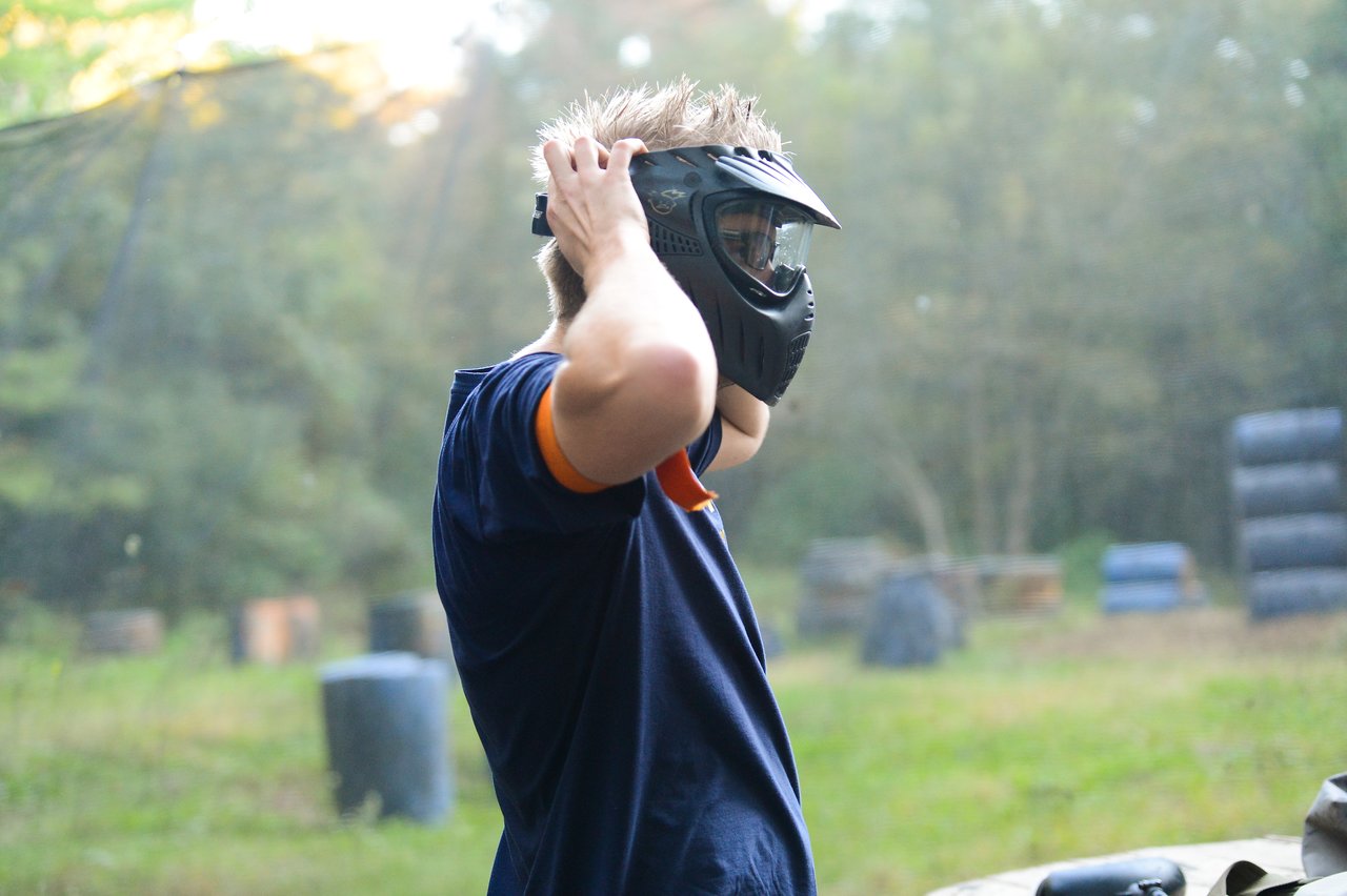 Person wearing a black paintball mask adjusts the straps while standing outdoors on a paintball field.