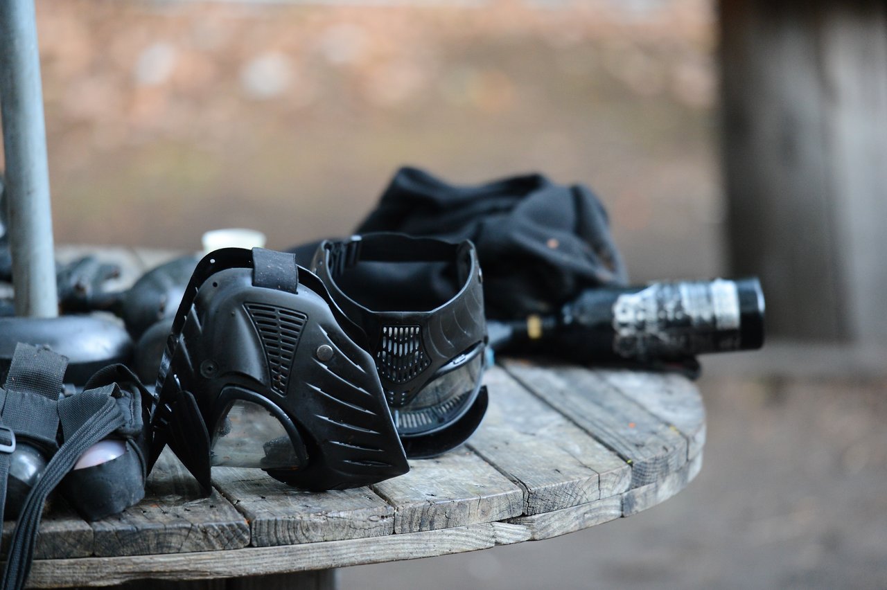 A black paintball mask and gear rest on a wooden table outdoors.