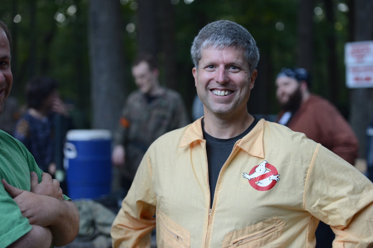 A smiling person in a Ghostbusters-themed jumpsuit stands outdoors with others in the background.