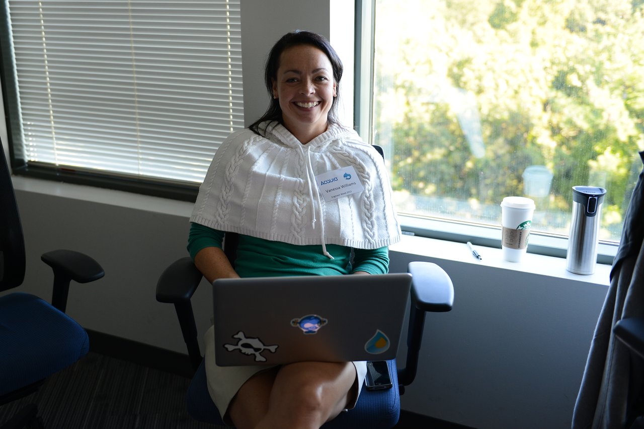 A person sits in an office chair, smiling while using a laptop, with a name tag on their sweater.