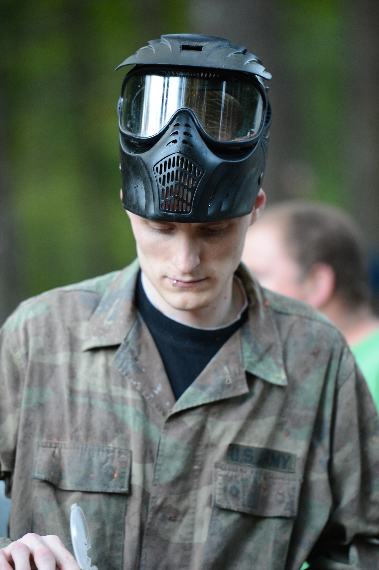 A person wearing a paintball mask and camouflage jacket looks down while handling equipment.
