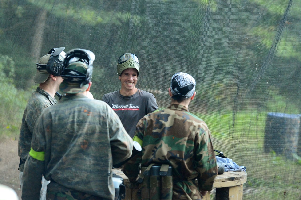 A group of people in paintball gear stand together, with one person smiling and wearing a helmet.