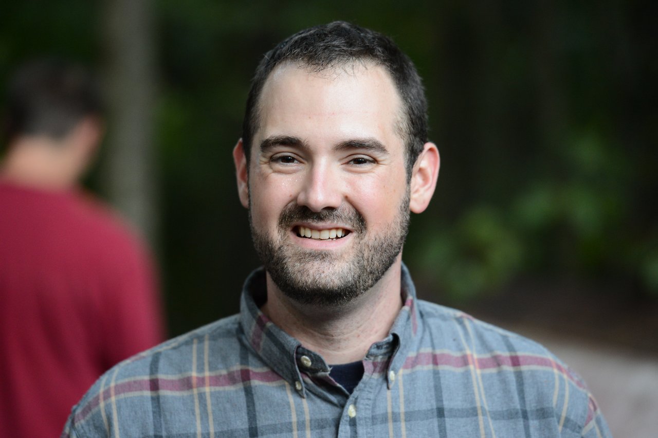 A man with a beard and plaid shirt smiles at the camera, with another person blurred in the background.