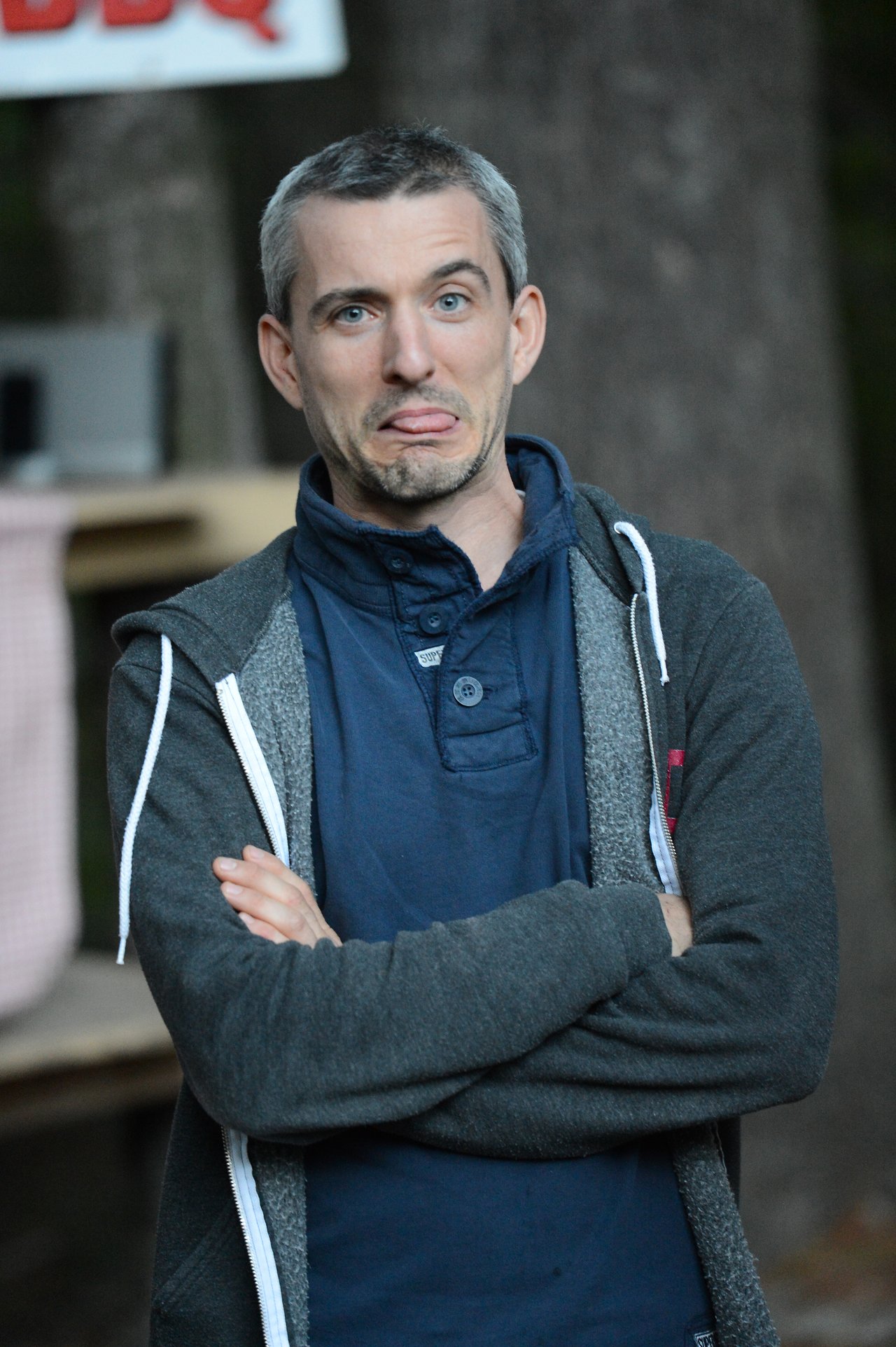 A person with short hair and a hoodie crosses their arms and makes a playful face, sticking out their tongue.