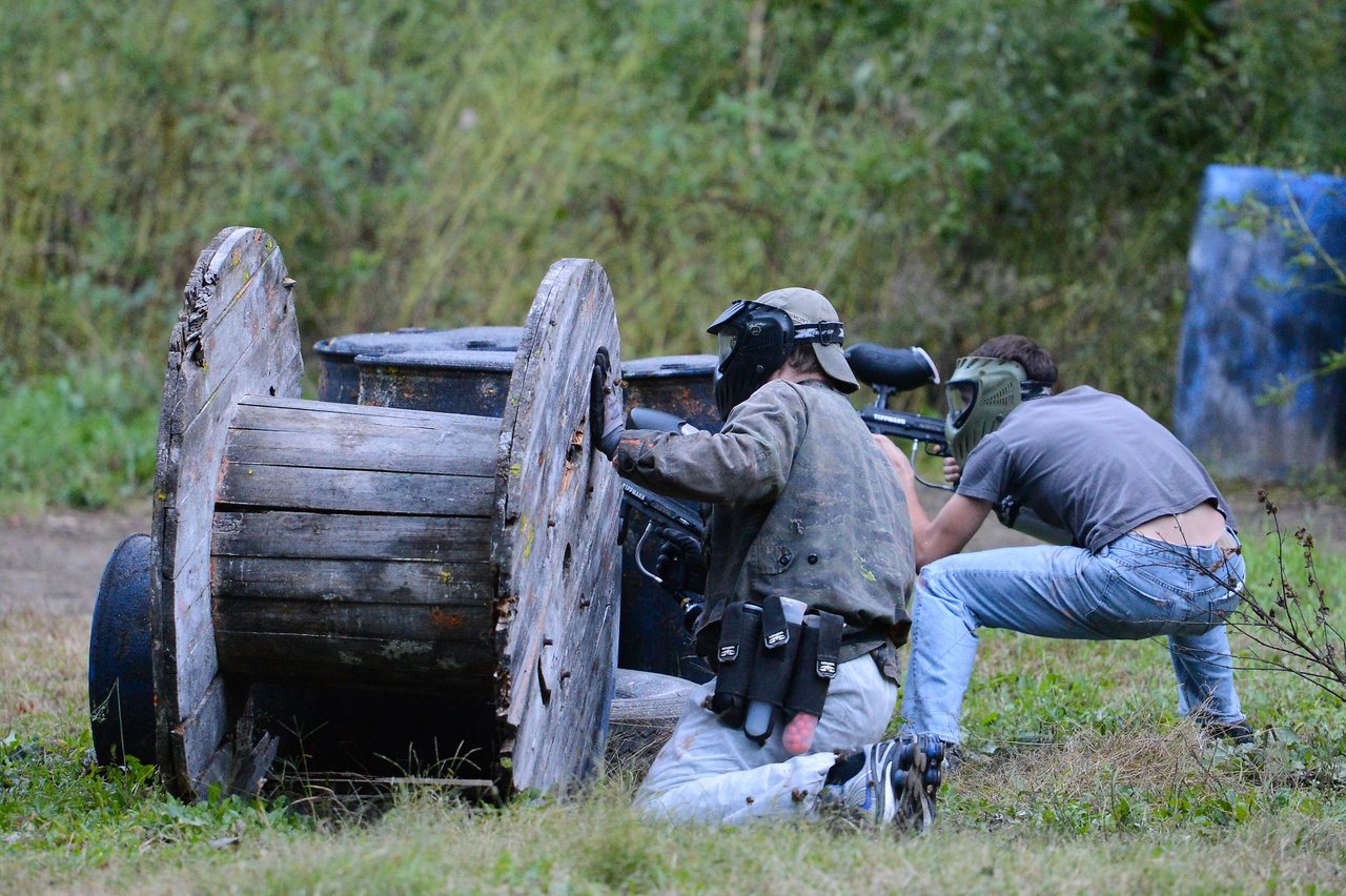 Two people wearing protective masks crouch behind a wooden barrier, aiming paintball guns during an outdoor game.