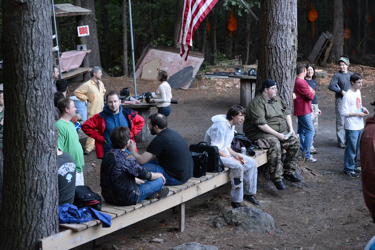 A group of people gathers outdoors, sitting and standing while talking and relaxing in a wooded area.