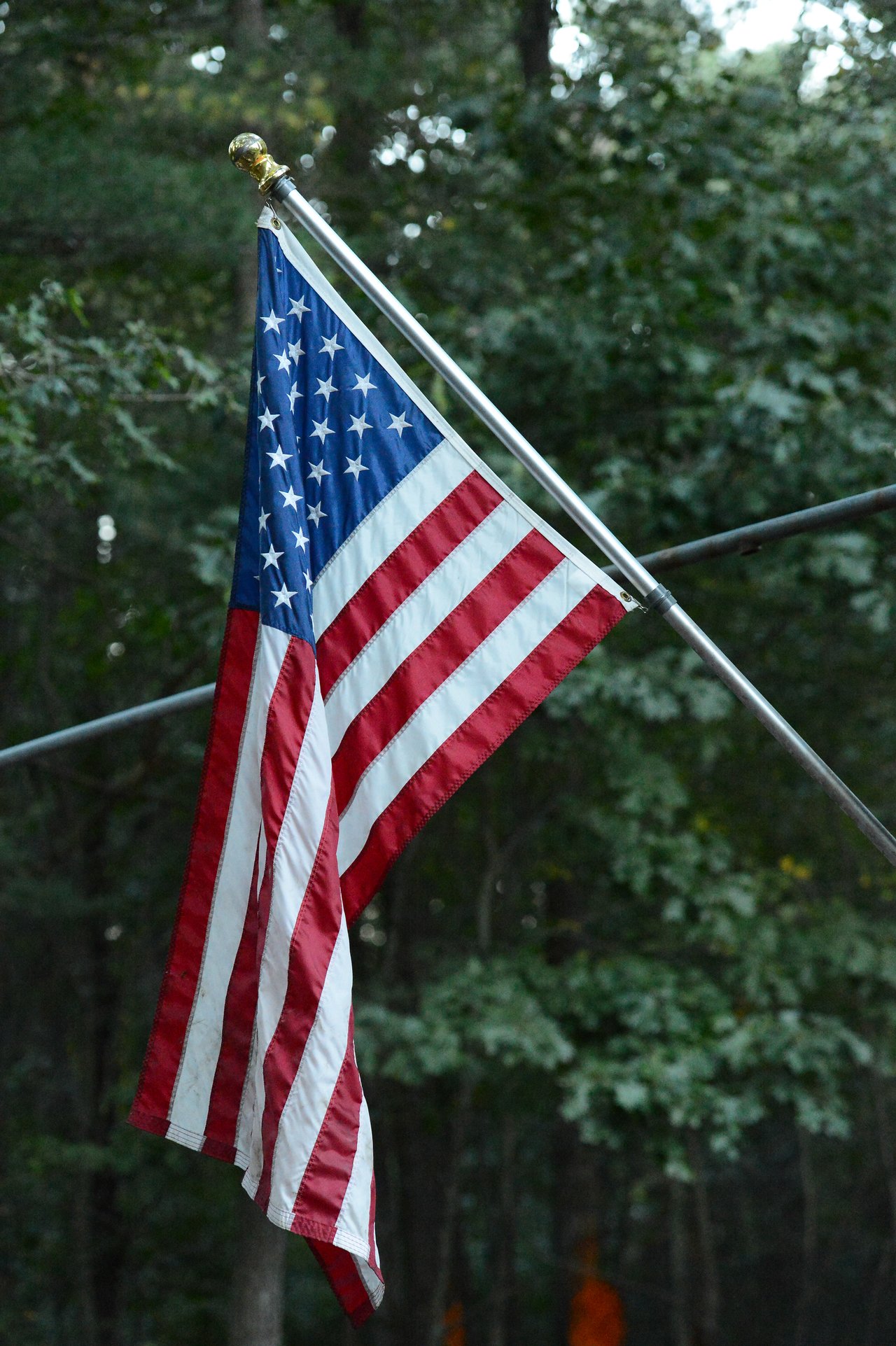 An American flag hangs from a pole, slightly folded, with a background of green trees.