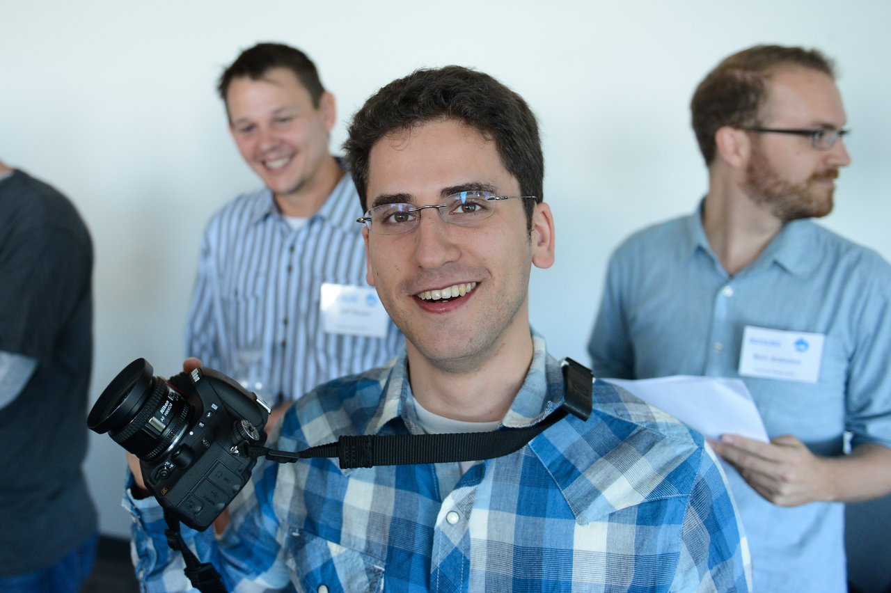 A smiling person in a blue plaid shirt holds a camera, with others in the background at an event.