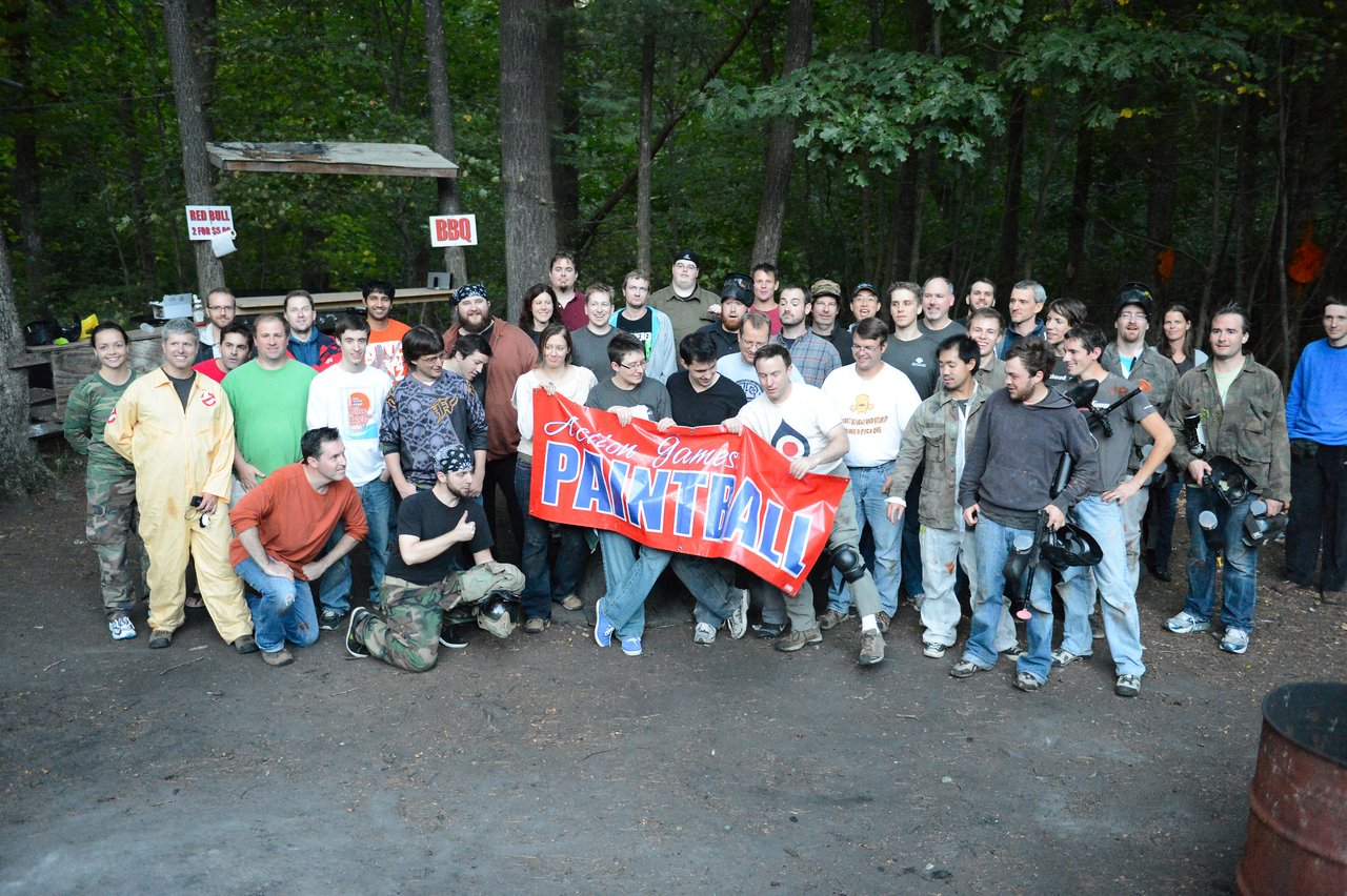 A large group of people poses outdoors, holding a "Paintball" banner, dressed in casual and paintball gear.