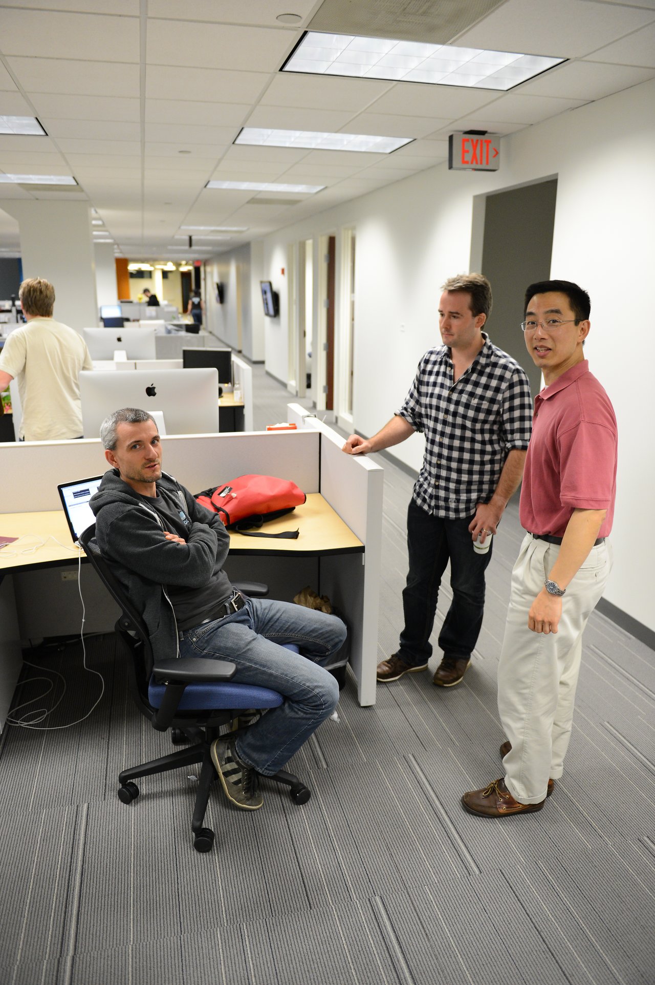 Three men in an office, one sitting at a desk while the other two stand and talk nearby.
