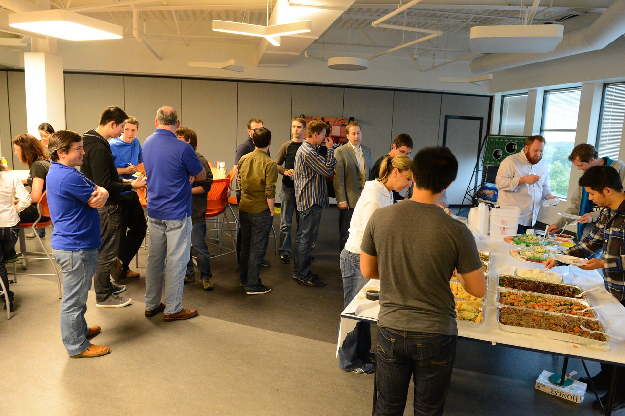 People gather in a room, chatting and serving themselves food from a buffet table during an event.