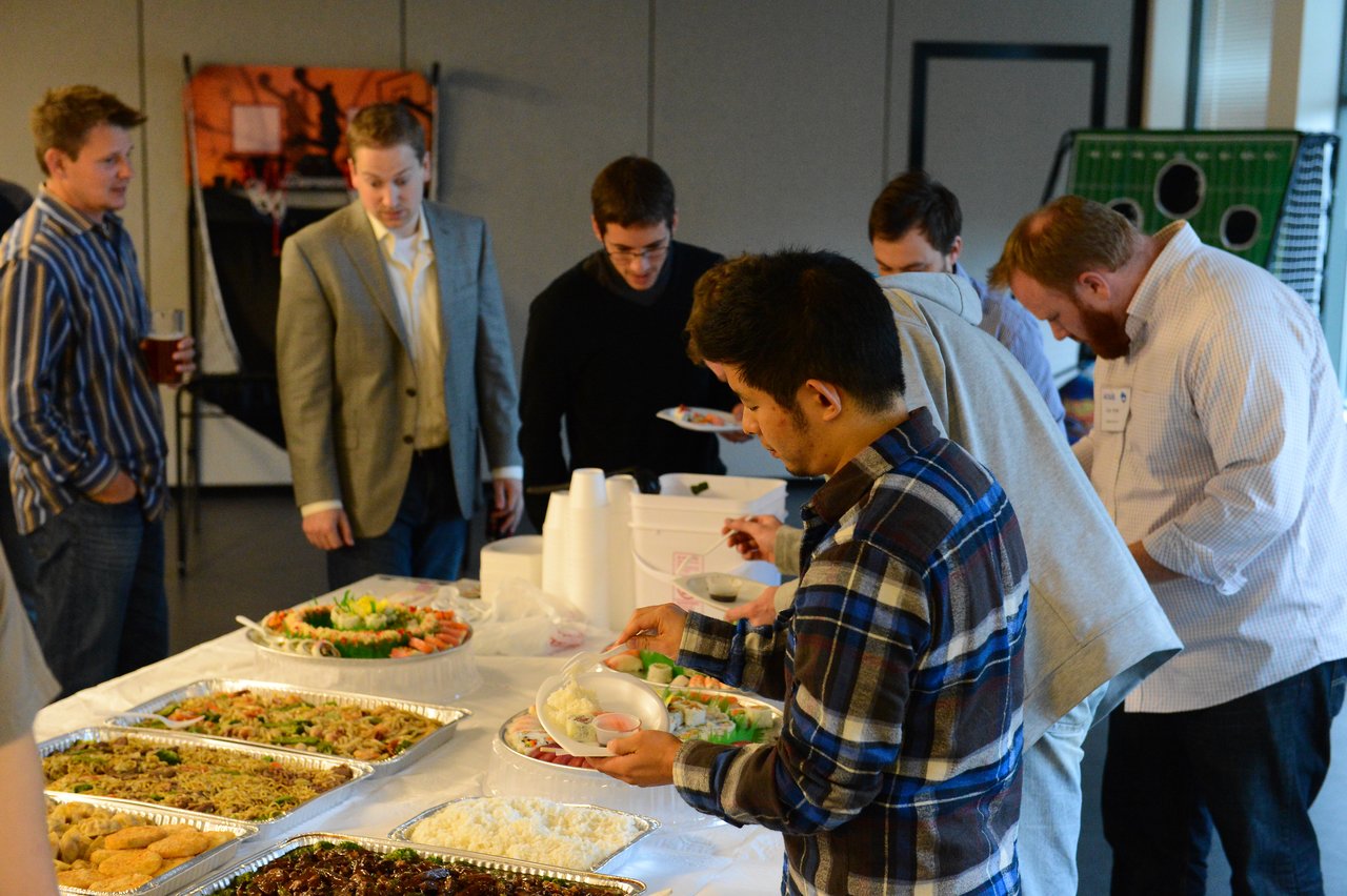 A group of people gathers around a buffet table, serving themselves food at an indoor event.